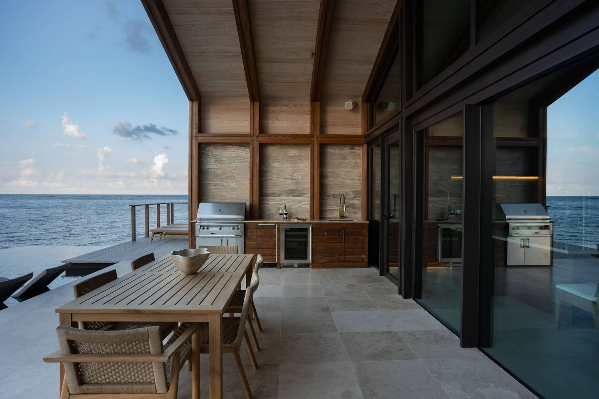 A modern outdoor patio at Ocean Villa, featuring a wooden dining table and chairs overlooking a vast ocean. The space boasts built-in grills and cabinetry beneath a covered structure with large glass doors, all set on a stone-tiled floor for the ultimate luxury experience.