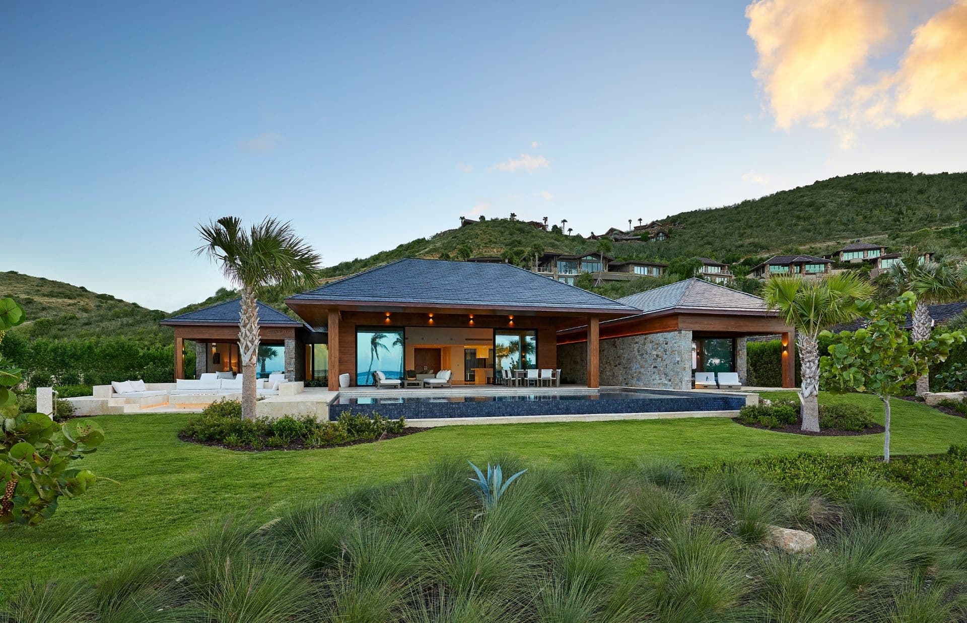 A modern, luxury house with large glass windows and a manicured lawn surrounded by tropical plants and palm trees. The house sits at the base of a green hill, with a partially cloudy sky in the background.