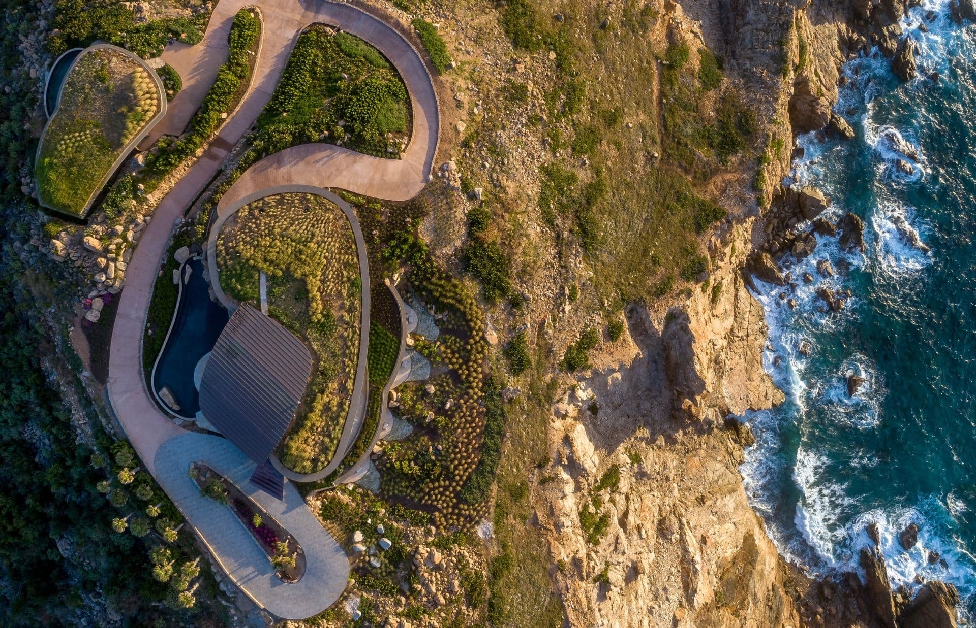 Aerial view of a modern, eco-friendly house with a green roof and curving pathways nestled on a rugged seaside cliff. Surrounded by natural vegetation, this Caribbean Developments property overlooks the ocean with waves crashing against the rocky shore below.
