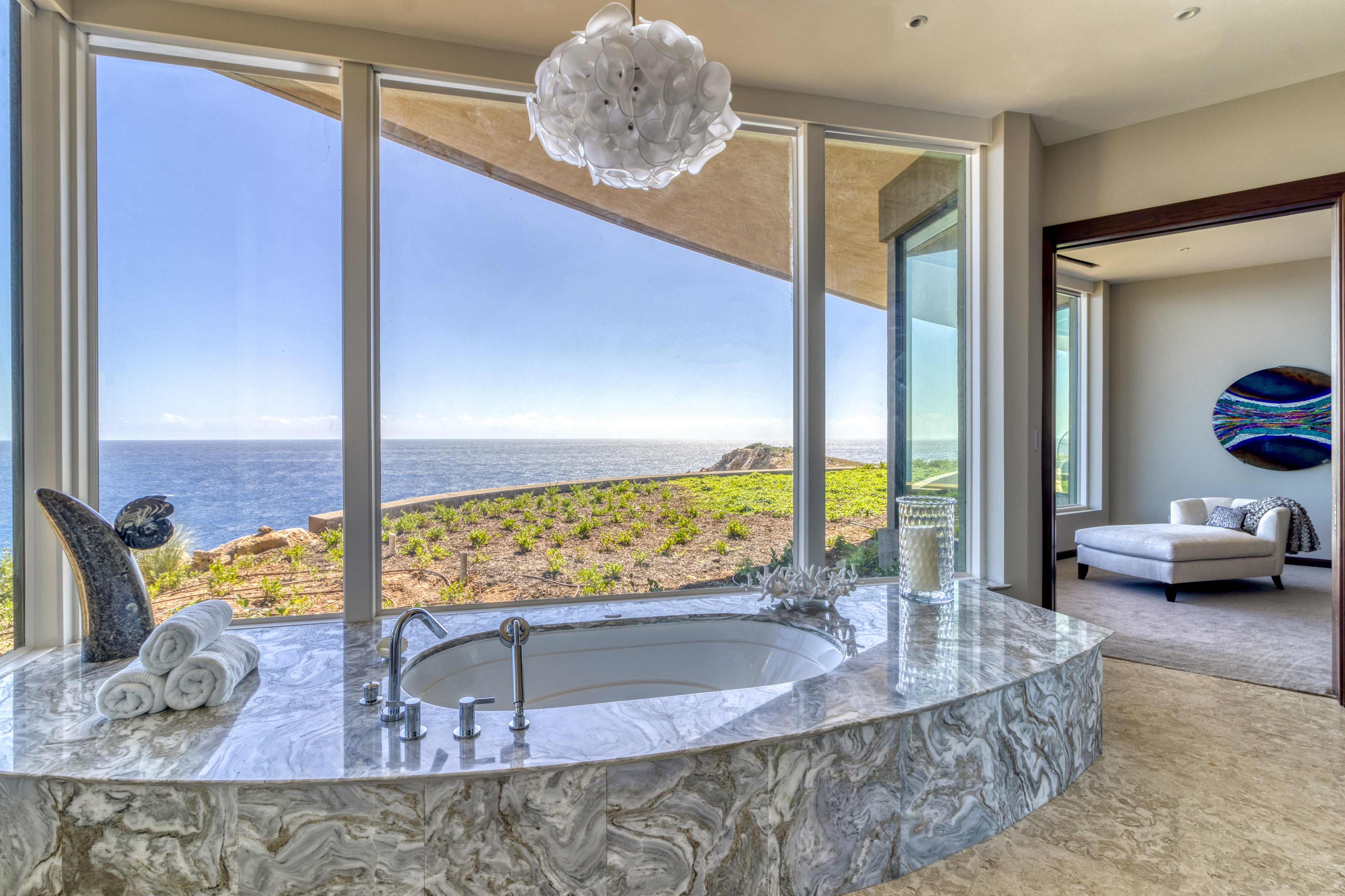 A luxurious Halo Villa bathroom features a large oval bathtub set in marble, positioned near floor-to-ceiling windows offering a stunning ocean view. The space includes elegant decor, a plush chair in an adjacent room, and a modern chandelier hanging from the ceiling.