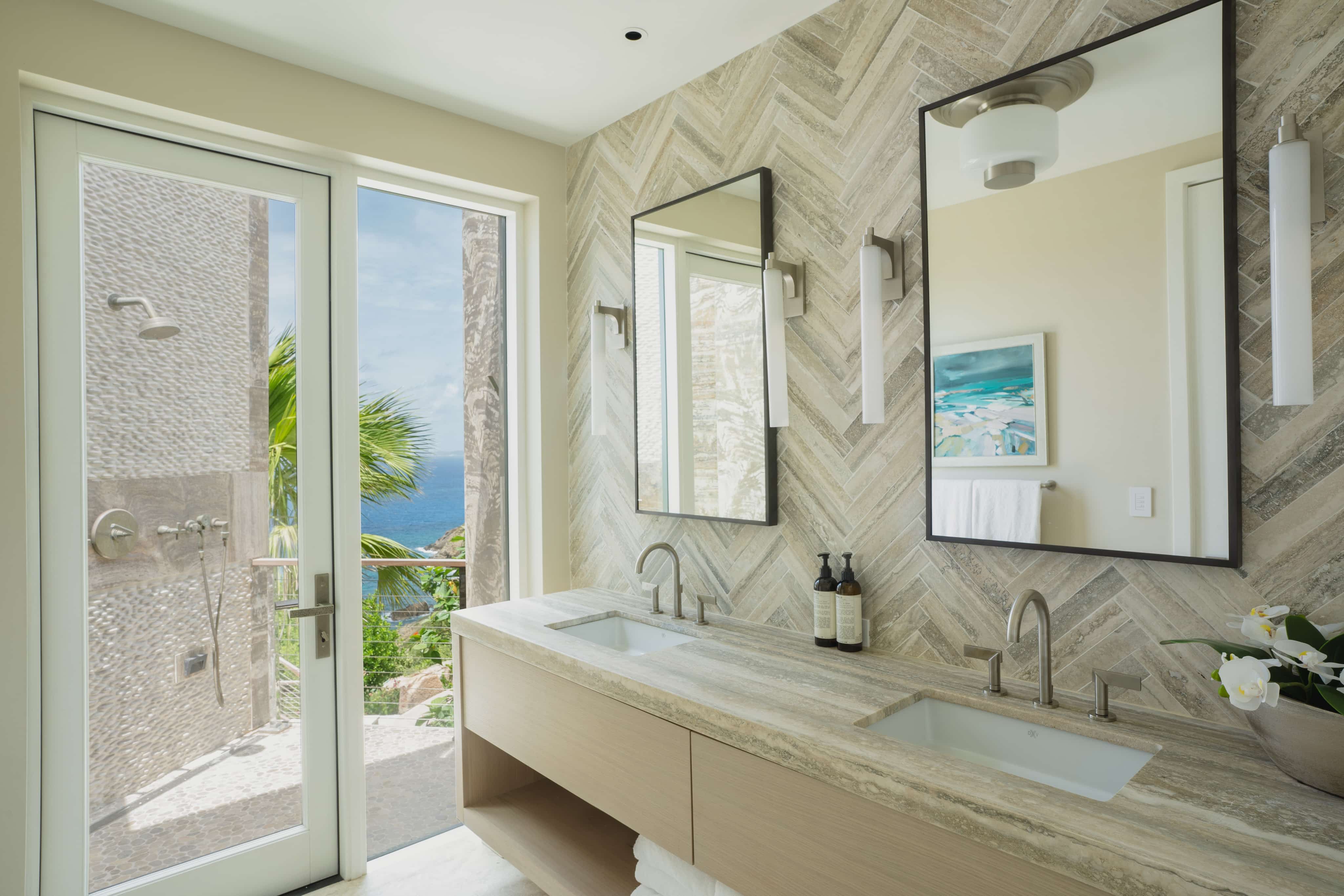 A modern bathroom in the luxurious Quintessa Villa features a large glass door opening to a scenic outdoor view with a glimpse of the ocean and tropical foliage. The interior includes a dual-sink vanity with a marble countertop, twin mirrors, sleek lighting fixtures, and decorative accents. Enjoy the ultimate vacation rental experience here.