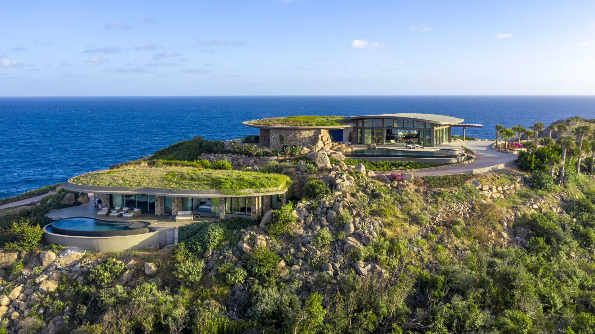 Aerial view of a luxury coastal home with two modern buildings featuring green roofs. The property is set on a rocky hill overlooking a clear blue ocean, surrounded by greenery and scenic natural landscape under a partly cloudy sky—a scene straight out of Boat International Magazine.