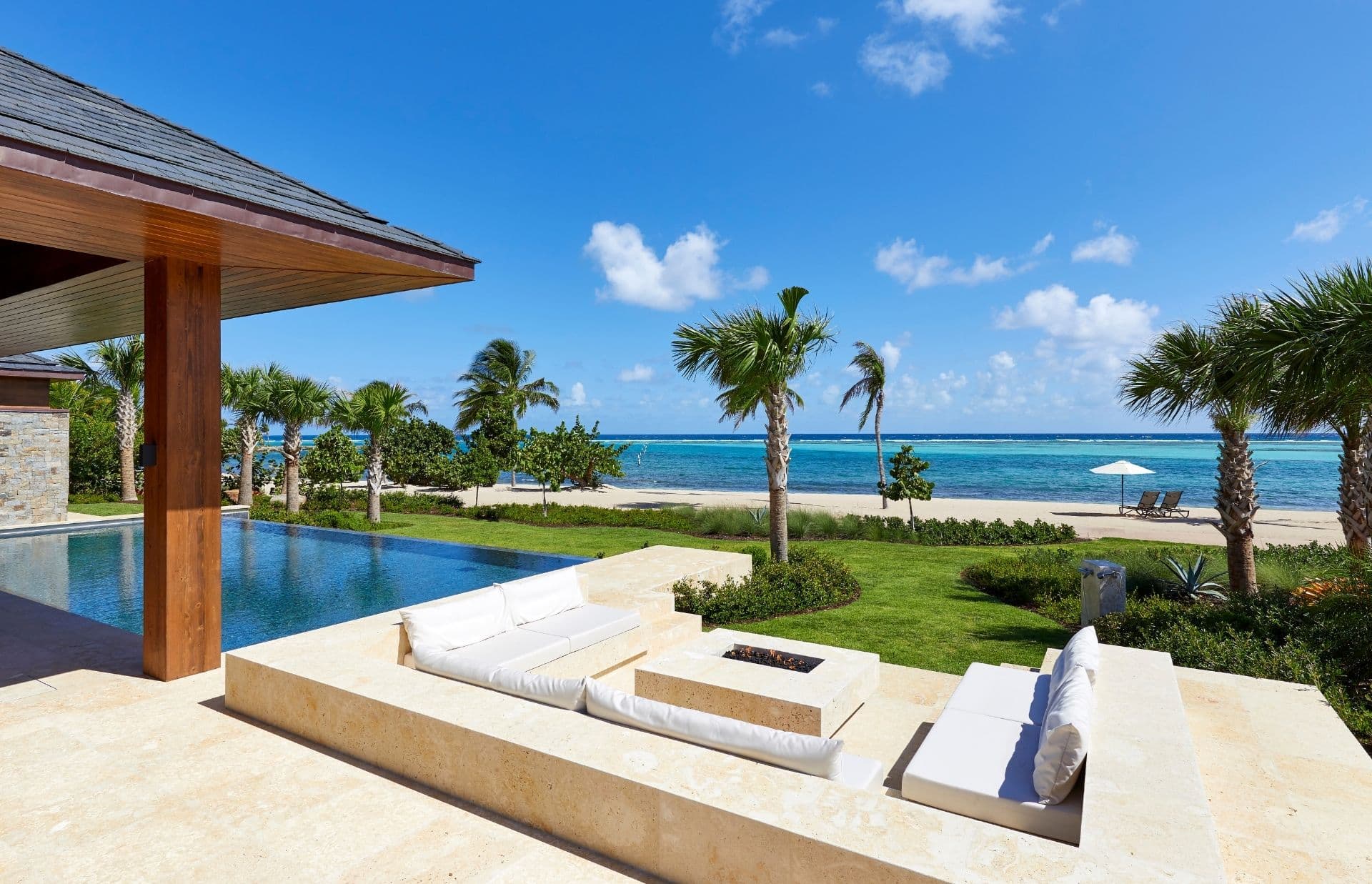 A luxurious beachfront patio overlooks a serene blue ocean. The patio features a modern infinity pool, white cushioned seating around a sunken fire pit, and palm trees providing a tropical ambiance. In the distance, sun loungers sit under a white umbrella on the sandy beach.