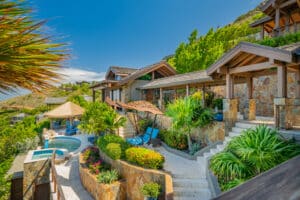 The image shows Poseidon's Perch Villa, a luxurious tropical retreat with a stone exterior nestled within lush greenery. There is a swimming pool with lounge chairs and thatched umbrellas, surrounded by beautifully landscaped gardens and a mountainous backdrop under a clear blue sky.