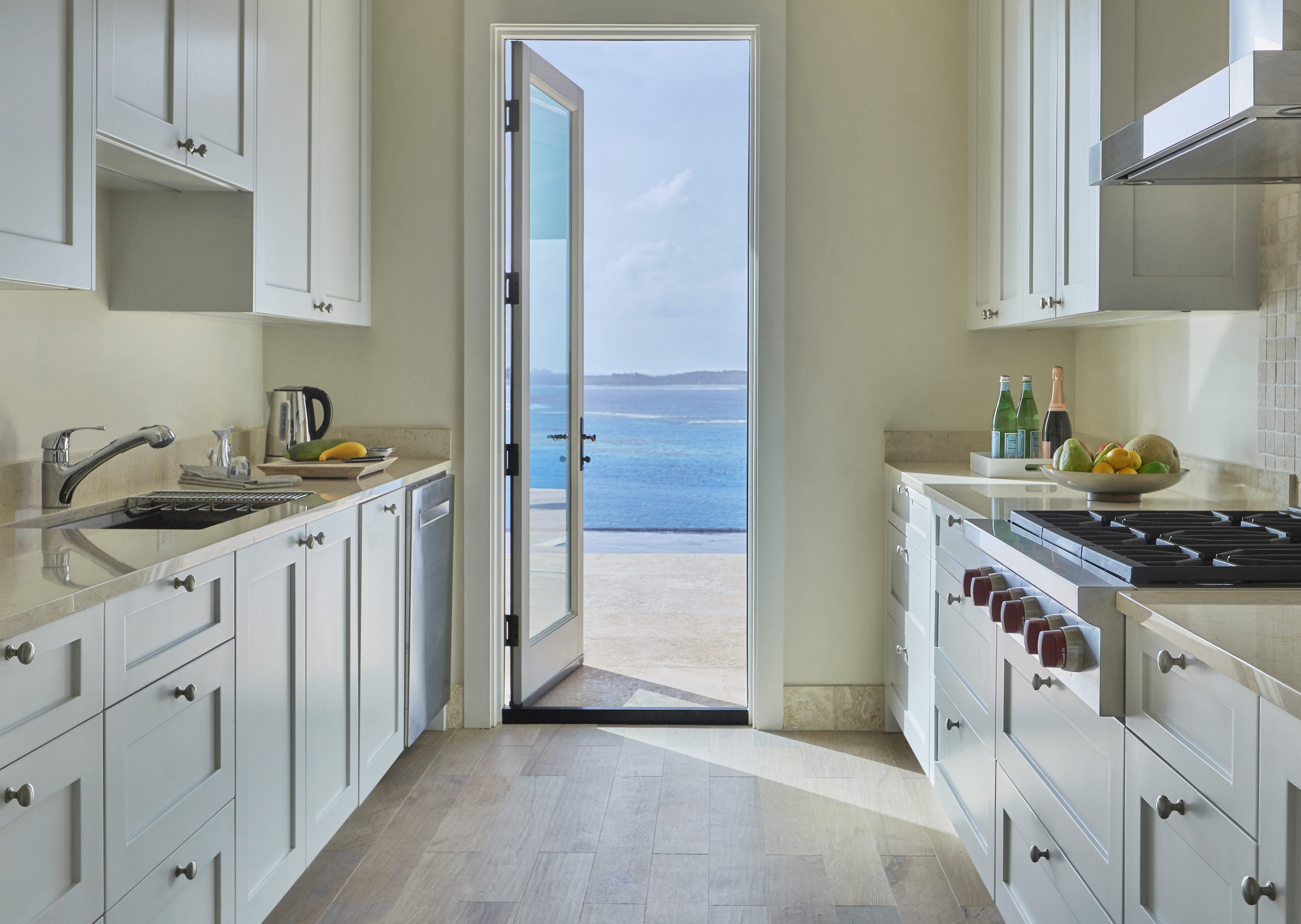 A modern kitchen with white cabinets, light beige countertops, and stainless steel appliances. The room features a door at the far end opening to a scenic view of the ocean, typical of Reef House. The countertops have a bowl of fruit, bottled water, and a few kitchen items.