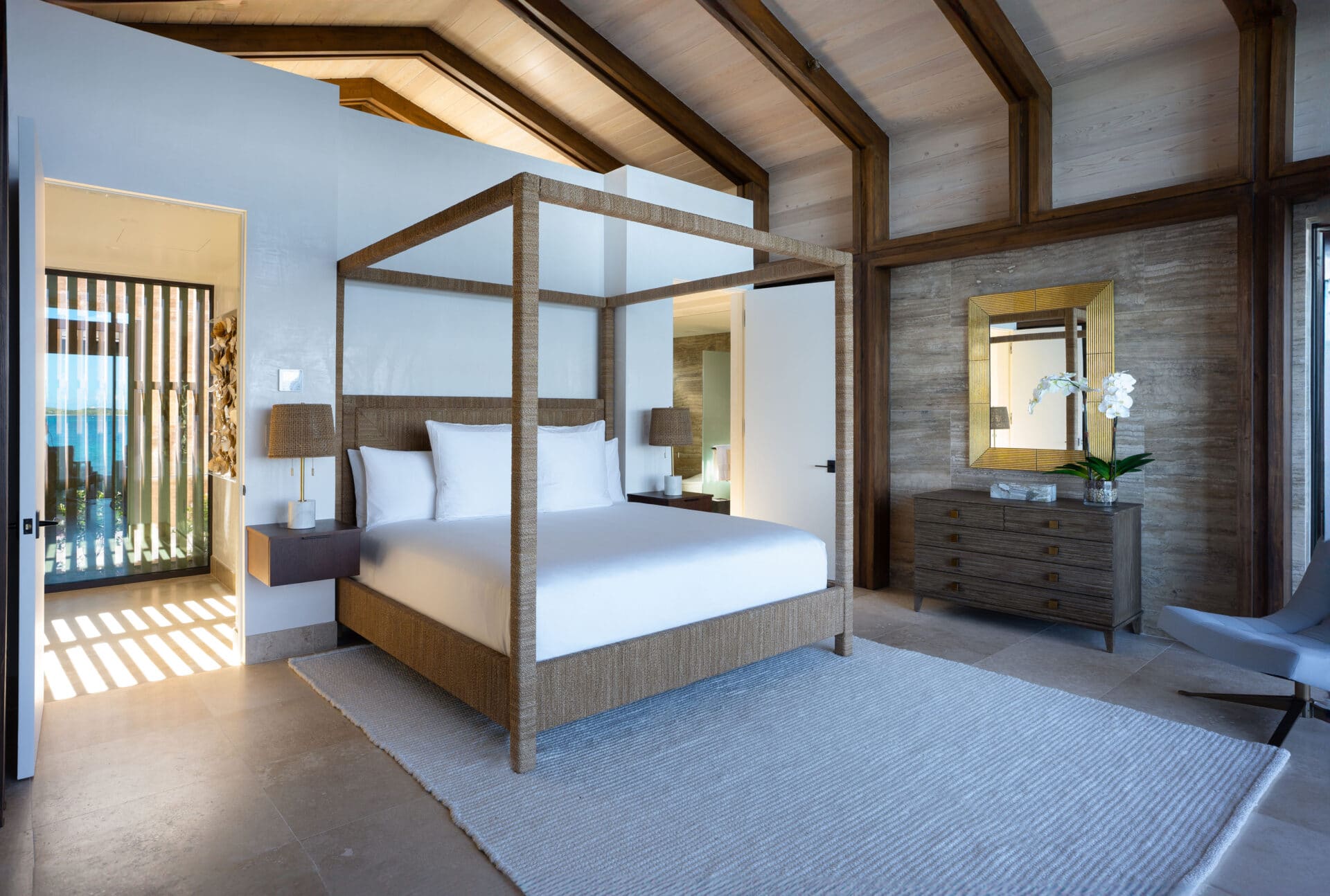 A modern Ocean Villa bedroom with a canopy bed featuring white linens, a light gray rug, and a wooden dresser with a mirror. The room has large windows letting in natural light, a small side table, and a decorative orchid plant.