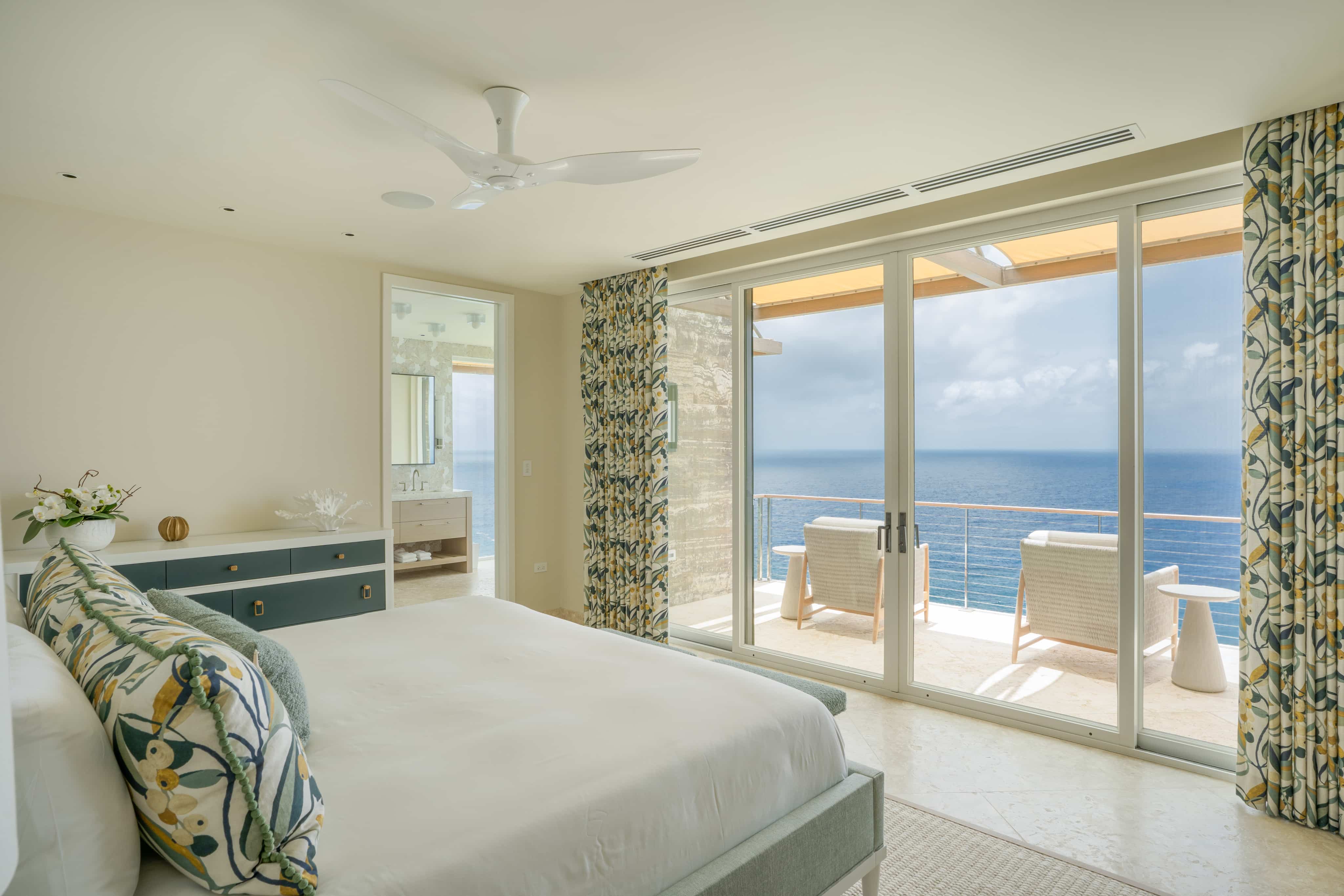 A serene bedroom in Villa Quintessa features a large bed with floral-patterned pillows and matching curtains. The room opens to a balcony through sliding glass doors, offering stunning ocean views. A stylish dresser and a ceiling fan complete the minimalist decor.