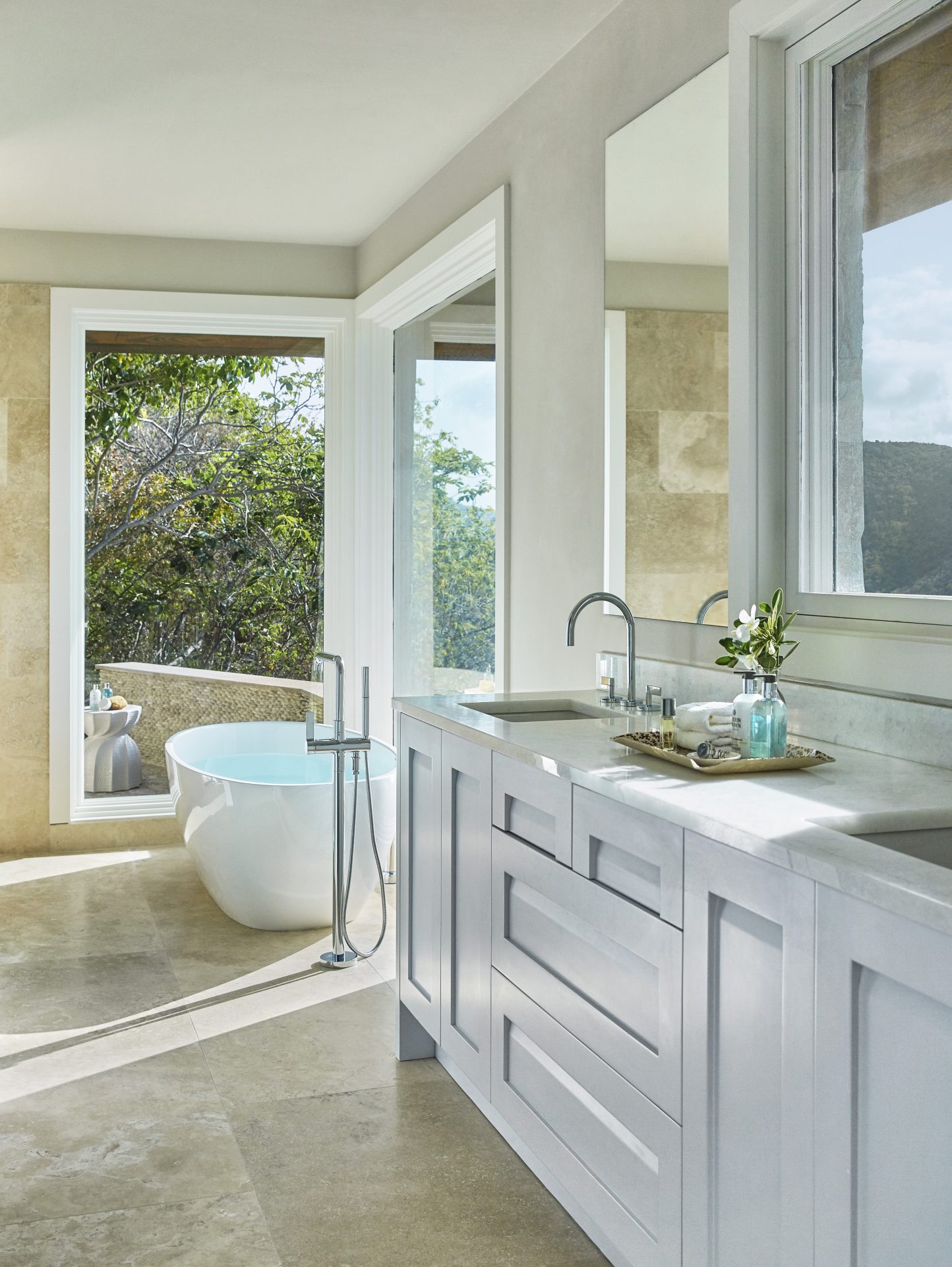A modern bathroom with light-colored cabinetry and countertops, a large mirror above the sink with twin faucets, and a tray with toiletries. A white freestanding bathtub is positioned by large windows overlooking a lush outdoor view, adding natural light to this serene house.