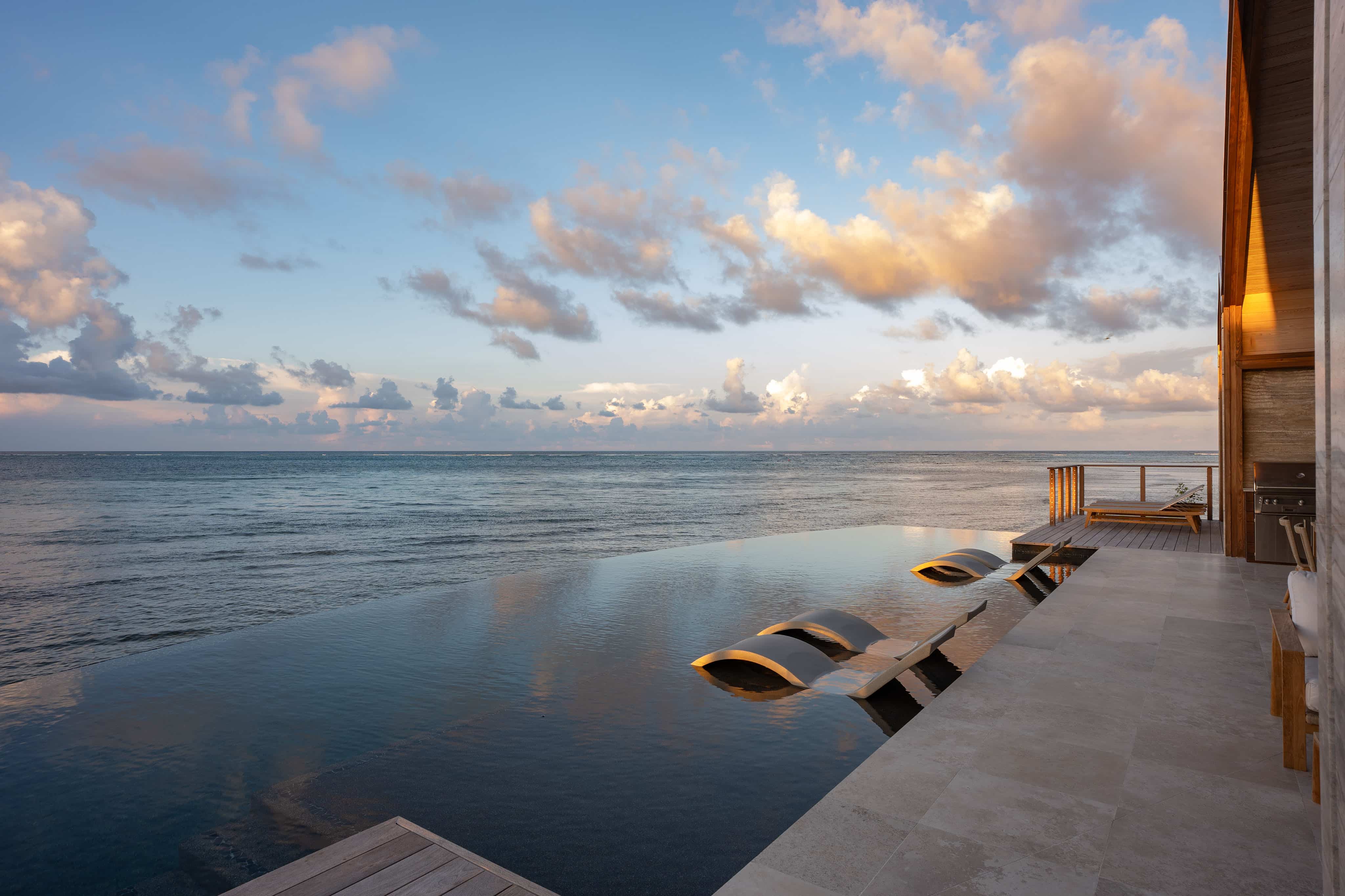 Perched beside a serene ocean view at sunset, Ocean Villa OV1 features an infinity pool with submerged lounge chairs that overlook the water. A modern wooden deck enhances the scene, creating a peaceful, luxurious atmosphere beneath fluffy clouds in the sky.