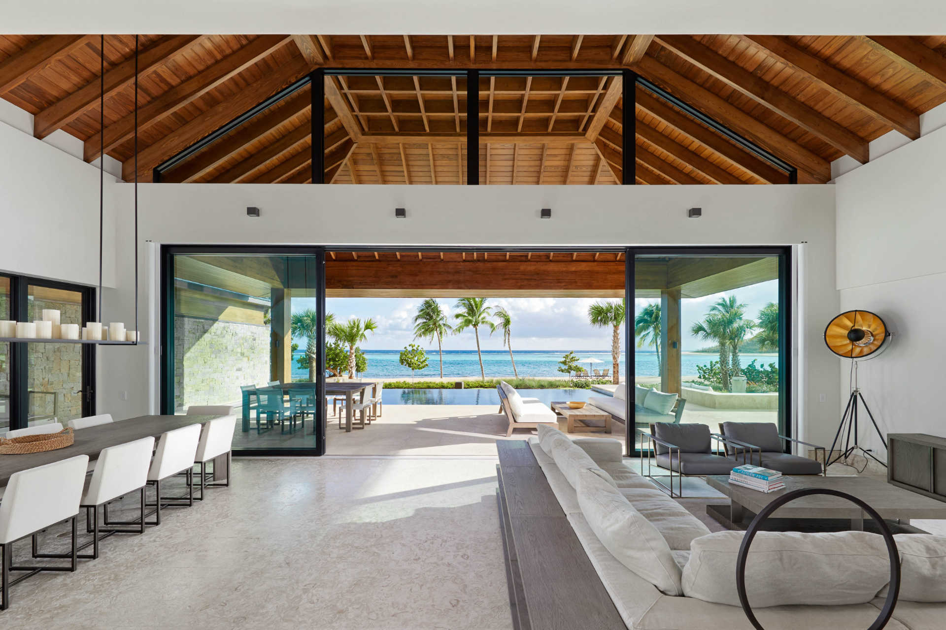 A spacious, modern living area in a stunning villa boasts a high wooden ceiling and large glass doors opening to a patio with outdoor furniture. The scene overlooks a beach with palm trees and a blue ocean. To the left, there is an elegant dining area with white chairs.