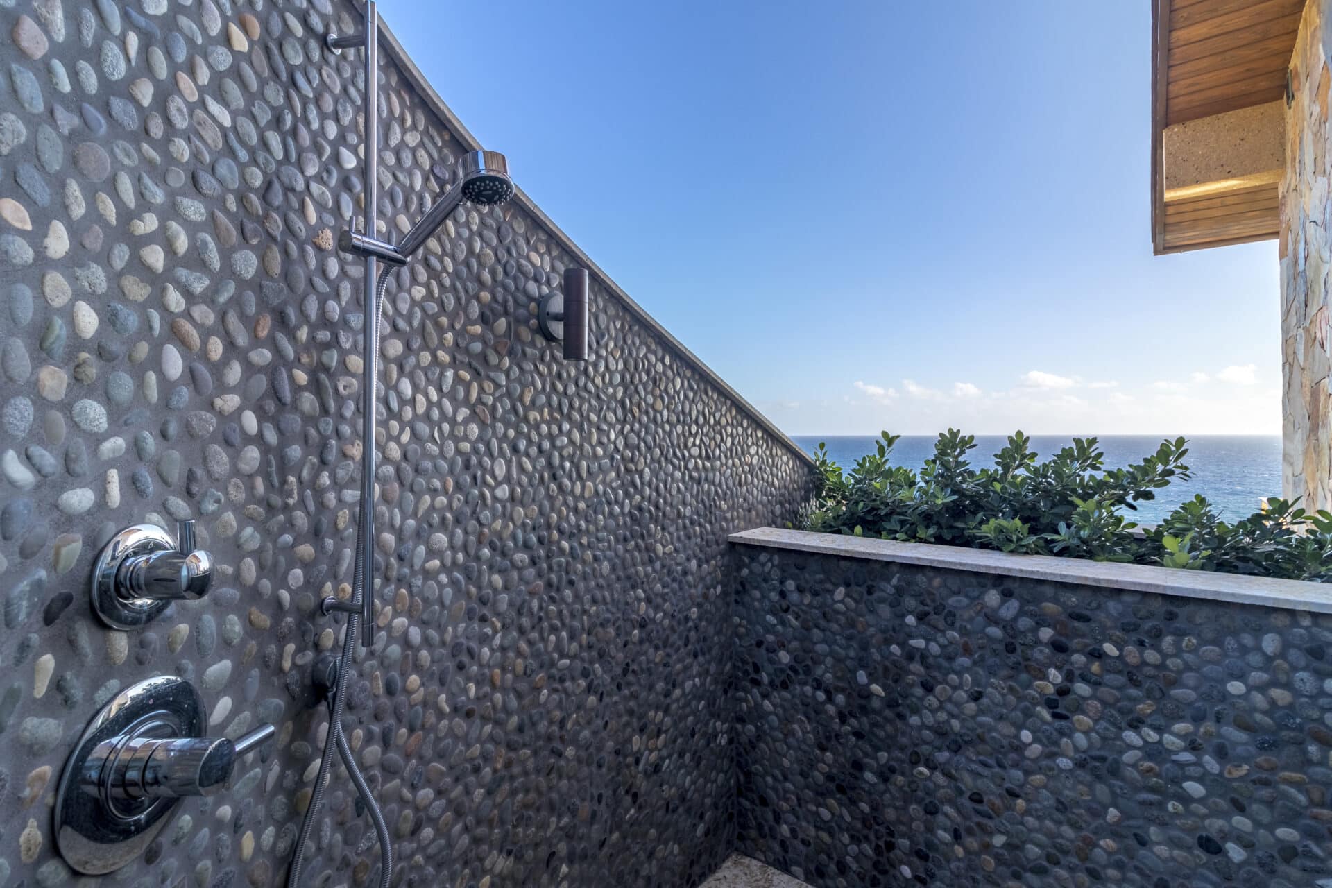Outdoor stone shower with modern metal fixtures at Wild Side Villa, overlooking a scenic ocean view under a clear blue sky. Lush green plants and a stone building edge partially visible on the right side.