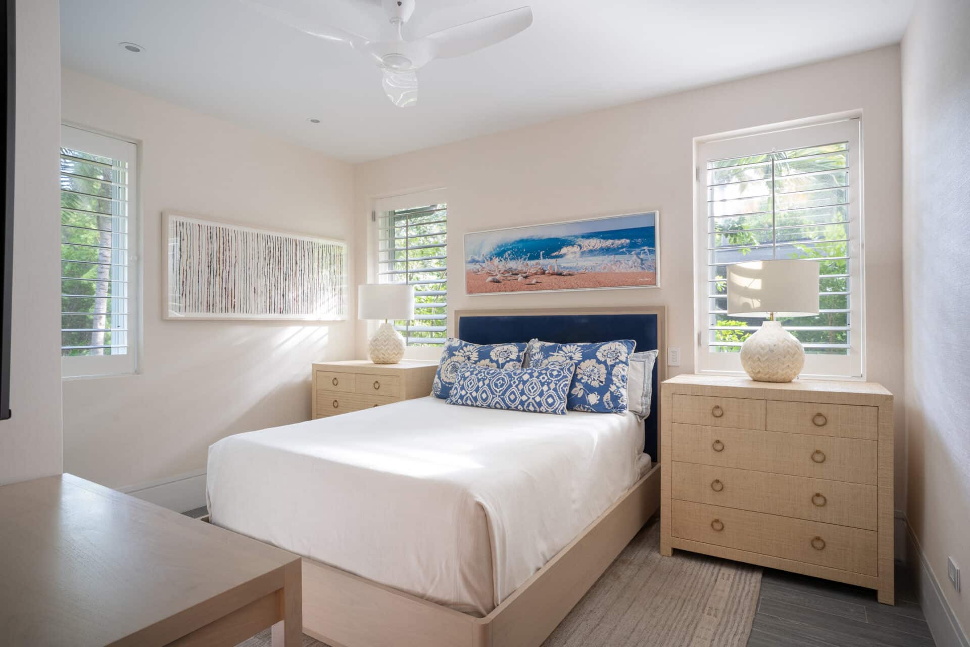 A bright bedroom in Nonamé Villa features a queen-sized bed centered between two wooden nightstands, each with a spherical lamp. The bed is adorned with white linens and blue, patterned pillows. Two windows with plantation shutters light the room, which is decorated with beach-themed wall art.