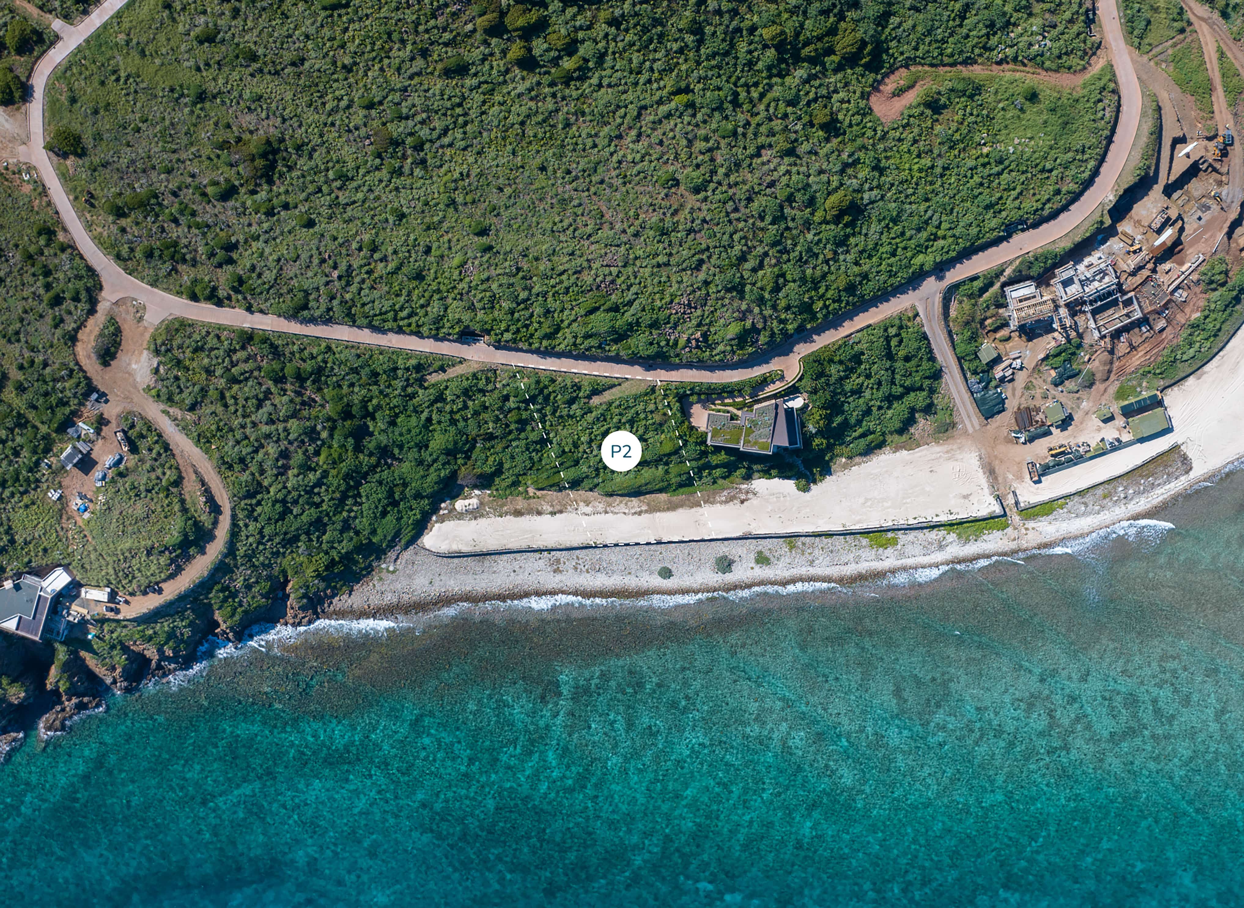Aerial view of a coastal area featuring clear turquoise waters, a sandy beach, and dense green vegetation. Near the shoreline, next to a small road on the peninsula, there's a homesite marked with a white circle labeled "P2".
