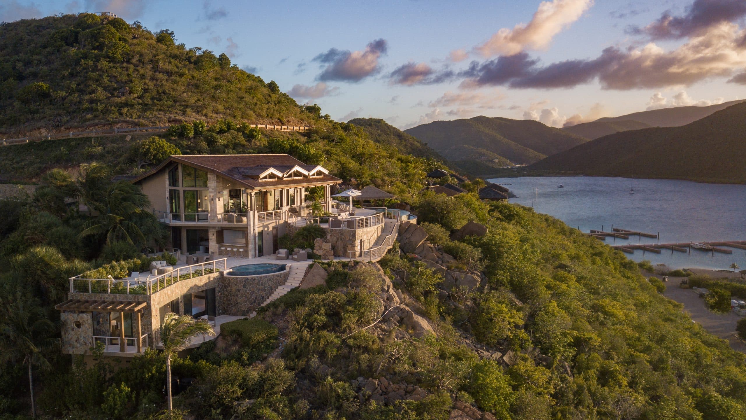 A luxurious hillside estate overlooking a serene bay with mountainous terrain in the background. The villa features multiple levels, expansive balconies, and a circular swimming pool, all bathed in the warm glow of the setting sun.