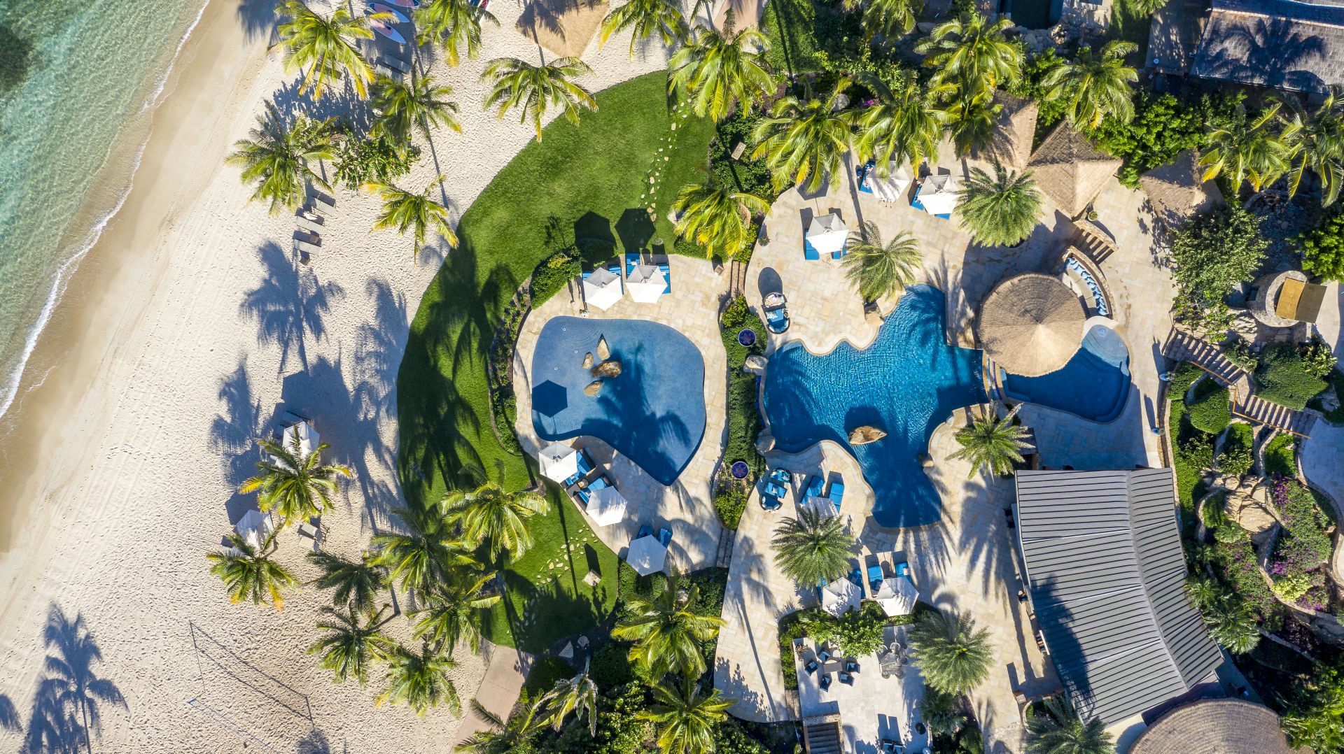 Aerial view of a tropical resort featuring two blue swimming pools surrounded by lush palm trees, green grass, and sandy beach adjacent to the ocean. Sun loungers and umbrellas are scattered around the pool areas, creating a serene vacation setting.