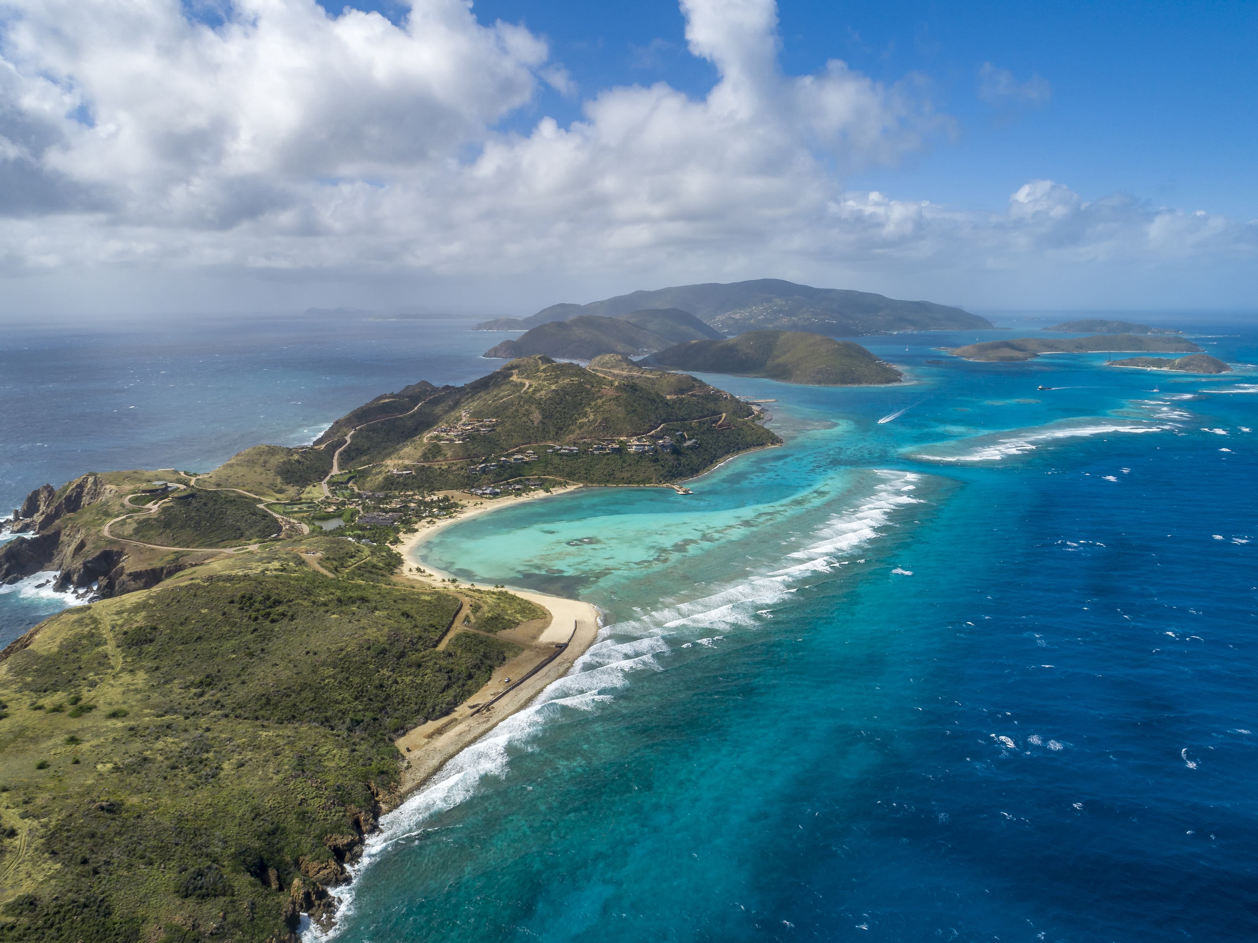 Aerial view of a tropical Island Homesite P2 with lush green hills, sandy beaches, and turquoise waters. The shoreline curves gently around the peninsula, bordered by coral reefs and shallow seas. The sky is partly cloudy, and other islands are visible in the distance across the ocean.