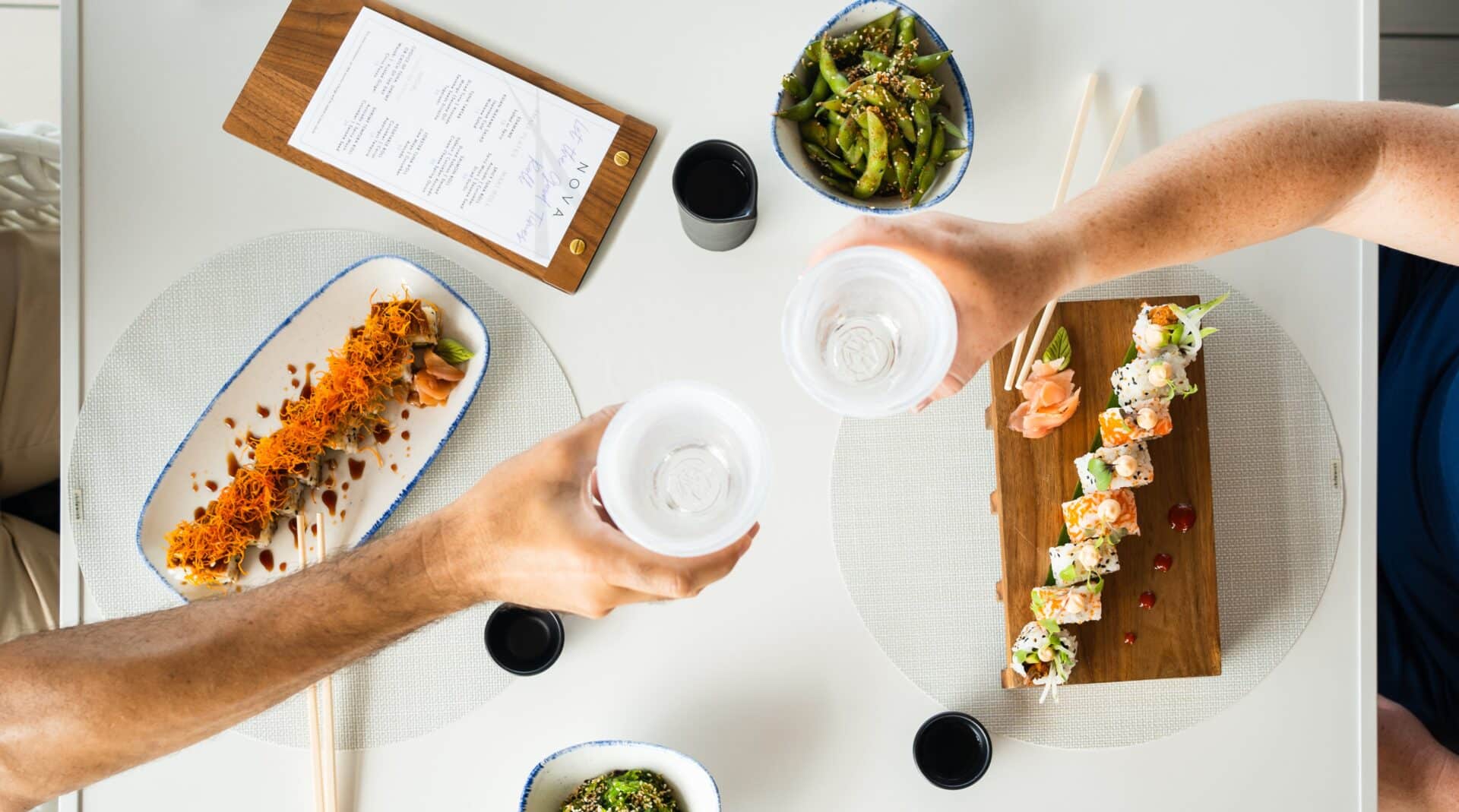 Two people clink glasses filled with water over a table set with sushi, including a dragon roll and a rainbow roll, edamame, a wooden menu, and dipping sauces on white place mats. This bright and inviting scene captures the essence of shared dining at Marina Village in the British Virgin Islands.