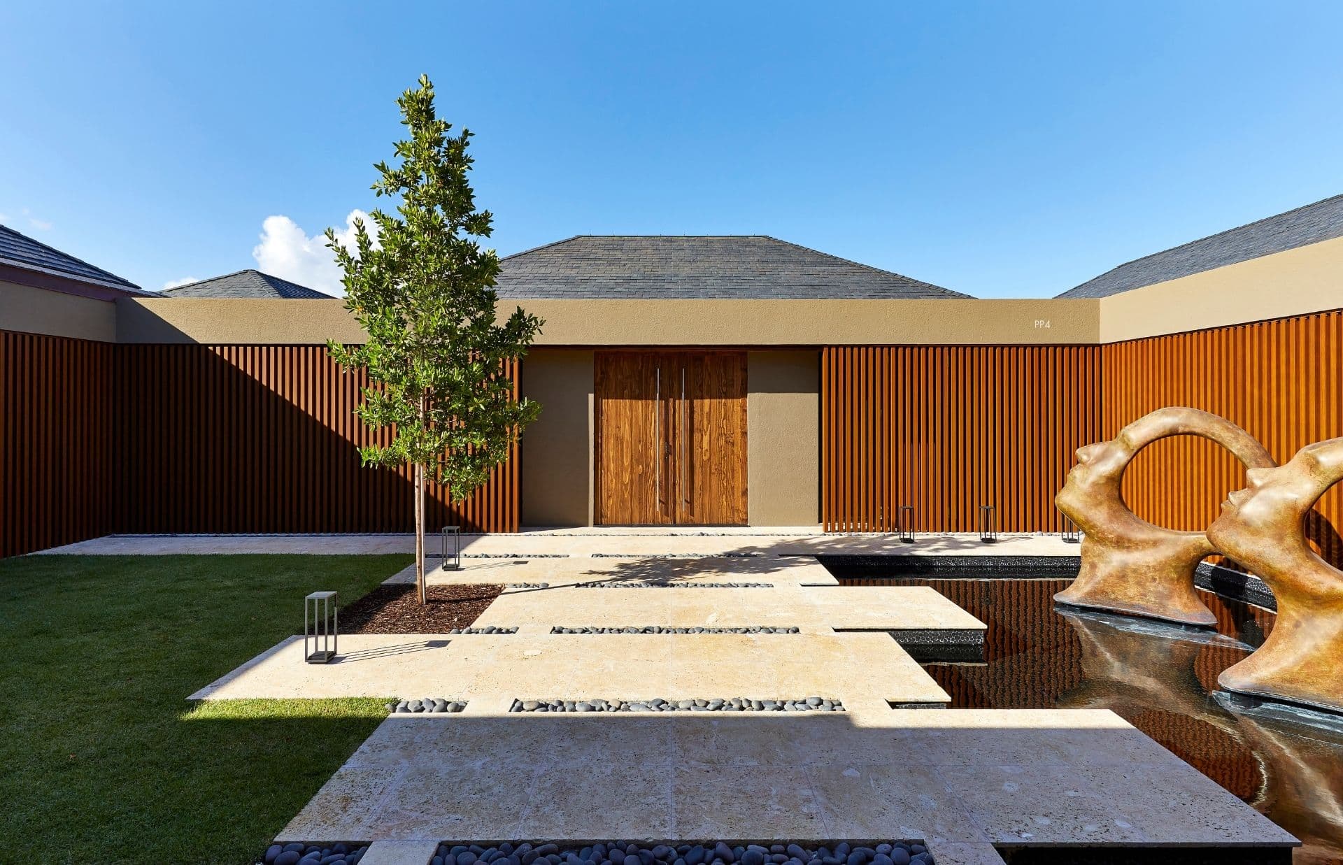 Modern courtyard with a single tree, a contemporary sculpture, and a water feature. The area includes a mix of grass, pebbled surfaces, and stone pathways leading to a wooden double-door entrance. Perfect for a family hideout, the walls have a wooden panel design. Sky is clear and sunny.