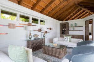 A modern villa's living room with a wooden ceiling and large windows. It features a white sofa, gray armchairs, a coffee table, and a console table with vases. A wall art installation resembling wings and built-in shelves with decorative items can be seen in the background.