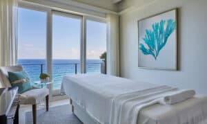 A serene spa room in Oil Nut Bay with a massage table facing floor-to-ceiling windows overlooking the ocean. A rolled towel rests on the table. The room is decorated with turquoise coral artwork and a small side table, perfect for a luxury escape in the British Virgin Islands.
