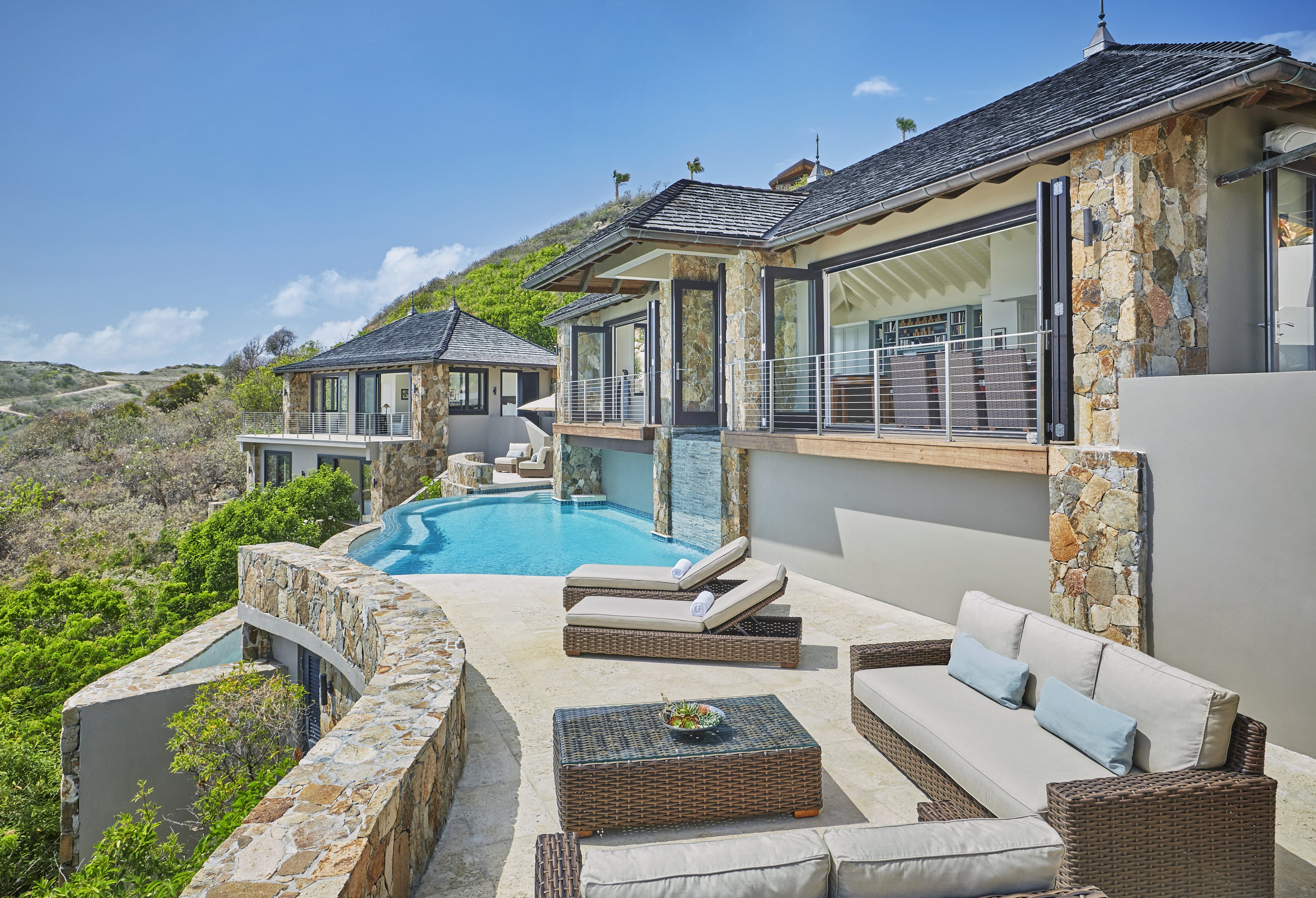 Cooke Lookout Villa is a luxurious hillside retreat with stone walls and an infinity pool that overlooks a lush green valley. The outdoor area includes sun loungers, a wicker coffee table, and a cushioned patio set under a clear blue sky. Adjoining rooms have large windows and sliding doors for breathtaking views.