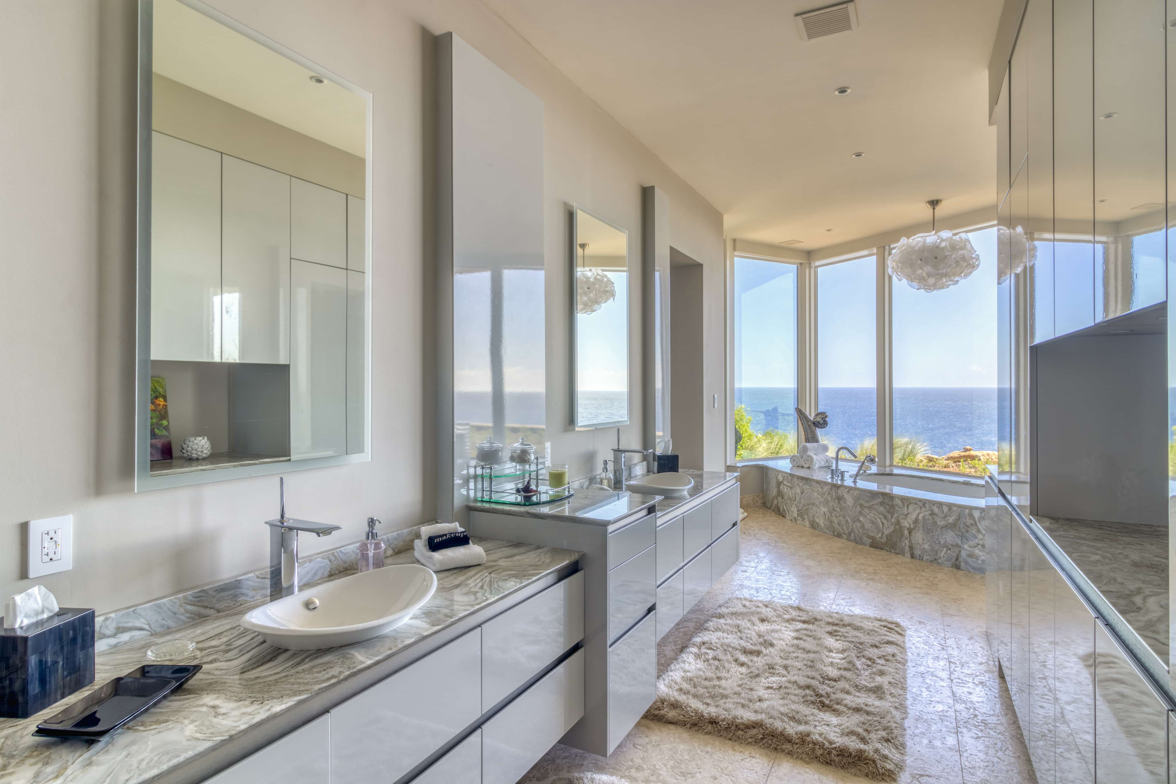A modern bathroom in Halo Villa features a marble countertop, dual sinks, wall-mounted mirrors, and minimalistic decor. A large window offers an ocean view. The room is bright with natural light, and next to the window sits a bathtub with a fluffy rug on the marble floor.