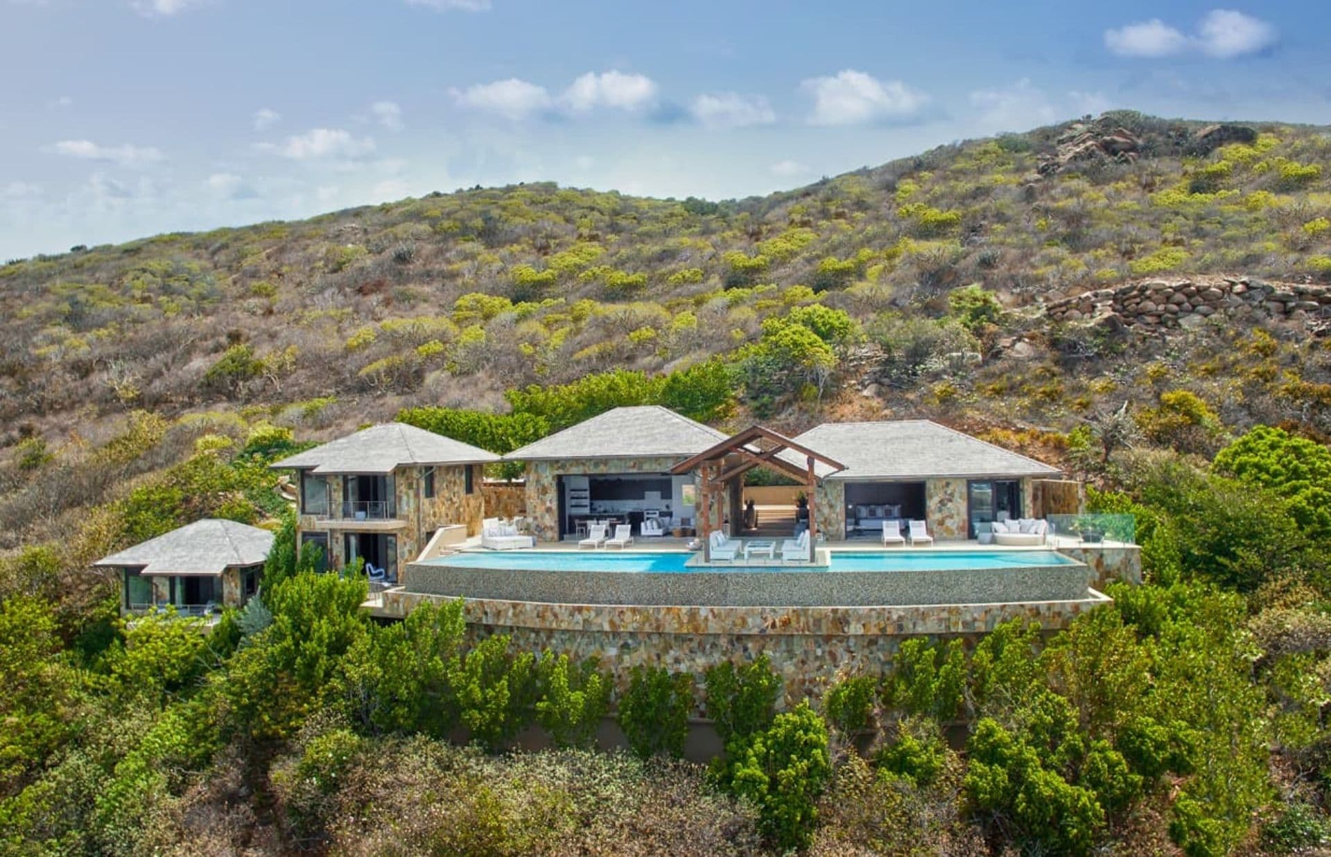 A luxurious villa featuring an infinity pool sits on a lush, green hillside under a partly cloudy sky in the British Virgin Islands. The villa, with its modern architectural elements and multiple structures connected by open spaces, offers breathtaking views and is surrounded by nature.