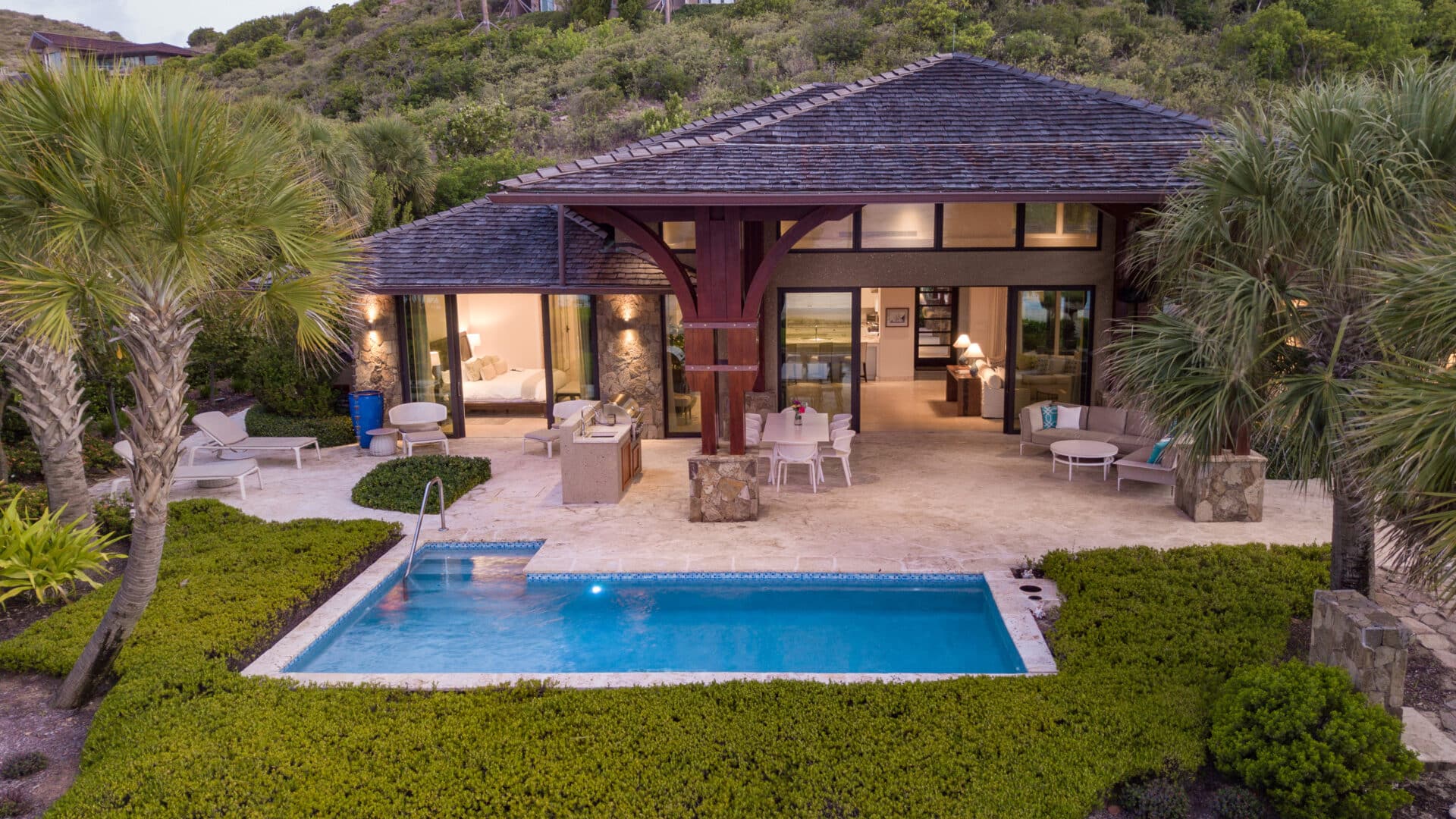 A luxurious, tropical Cheemaun villa with an inviting outdoor area featuring a small rectangular pool, surrounded by lush greenery and palm trees. The villa has large glass doors, an outdoor seating area, and appears cozy and well-lit from within.