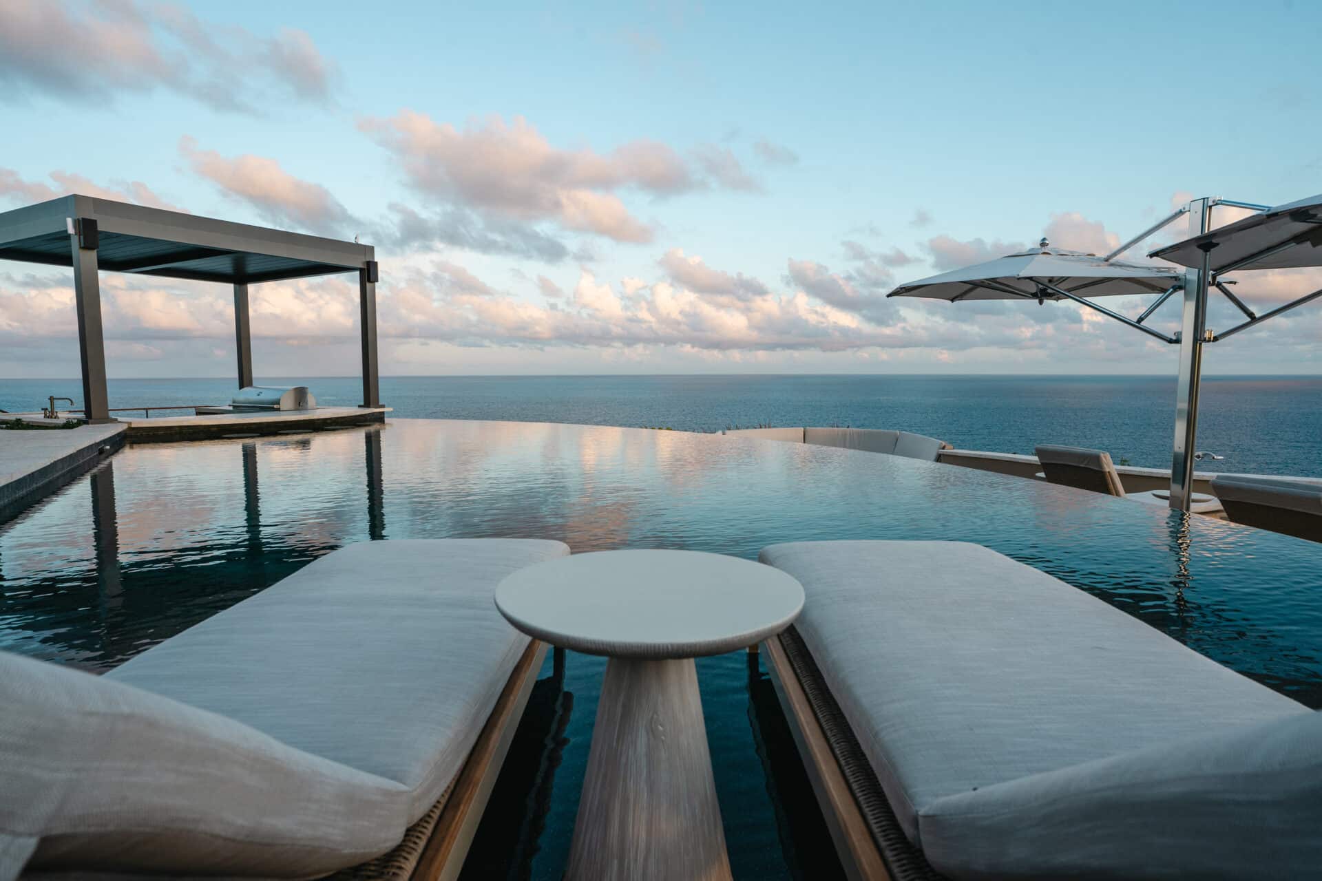Two cushioned lounge chairs are set beside an infinity pool overlooking the ocean at Quintessa Villa. A round table sits between the chairs, and large white umbrellas provide shade. A modern pergola structure is visible to the left, and the sky is filled with soft, scattered clouds.