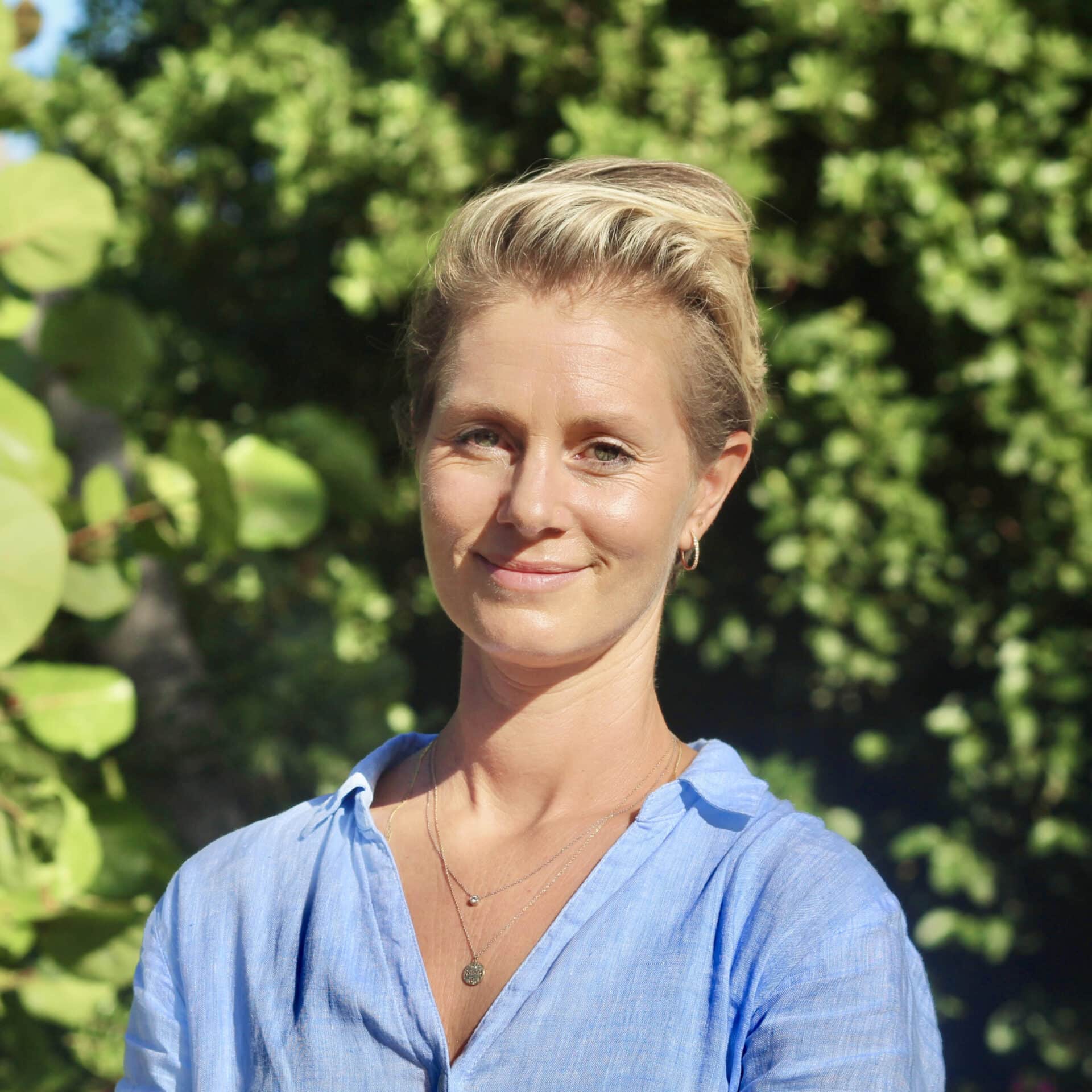 Emily Oakes, a woman with short blonde hair, stands outdoors in front of green foliage. She wears a light blue blouse and smiles gently at the camera. The bright lighting suggests a sunny day. Emily adorns two delicate necklaces and small hoop earrings.