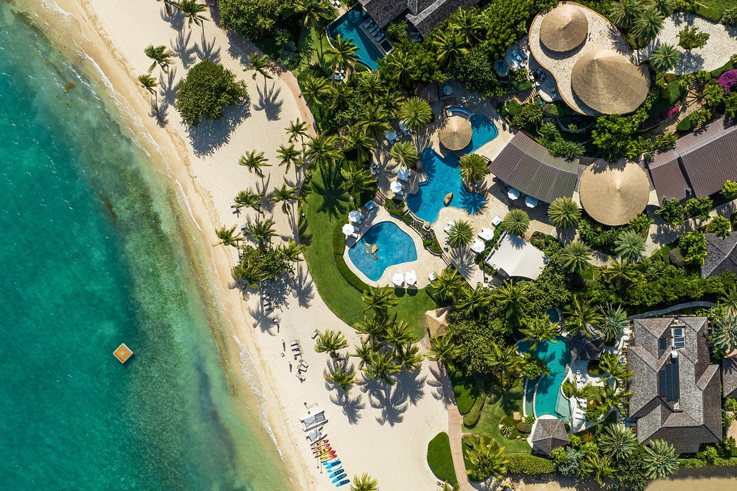 Aerial view of a tropical beach resort featuring lush greenery, several pools, and thatched-roof buildings. Sun loungers and colorful kayaks are lined up on the white sandy beach next to the vibrant turquoise sea. The Beach Club Pavilion at Oil Nut Bay completes this idyllic paradise.