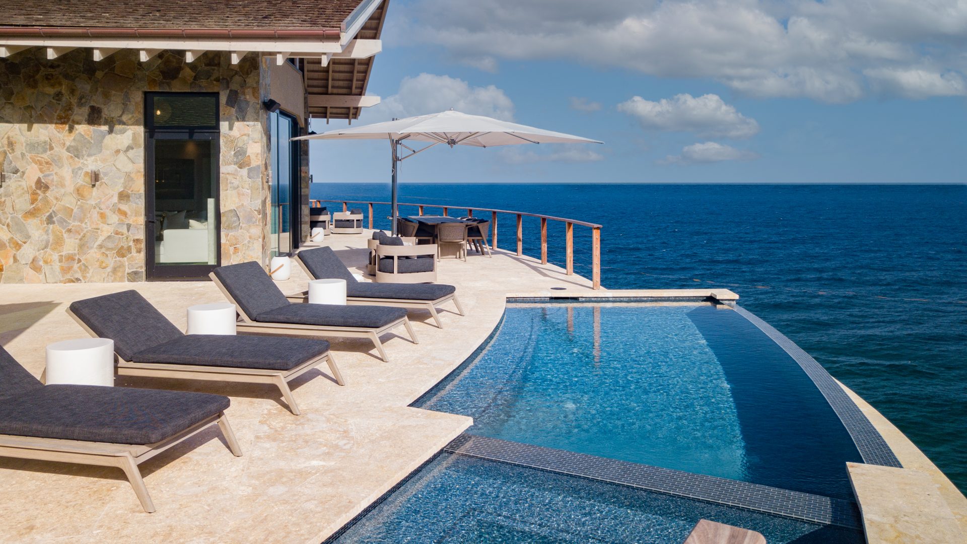 Luxury villa featuring a stone facade, an infinity pool, and four lounge chairs on a sun-drenched terrace overlooking the ocean. An umbrella shades a seating area with a table and chairs, creating a serene and picturesque setting under a partly cloudy sky where hummingbirds occasionally glide by.