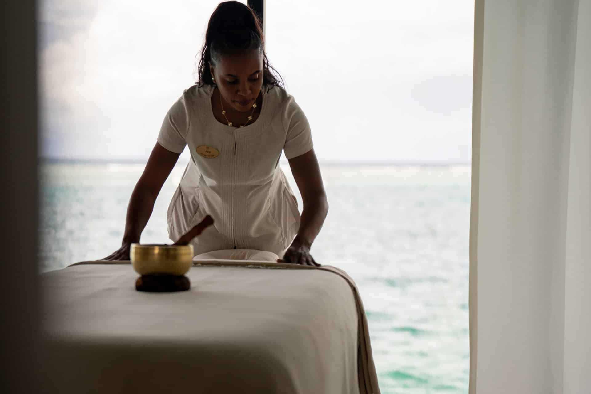A spa therapist in a white uniform prepares a massage table in a serene room with large windows overlooking a calm ocean at Oil Nut Bay spa, with a singing bowl placed nearby.