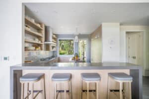 The modern kitchen at Nonamé Villa boasts a minimalist design, featuring a long, grey countertop with four light wood stools. Open shelving displays dishes and glassware, while a large window offers a view of greenery outside. The space is bright with natural light and clean lines.