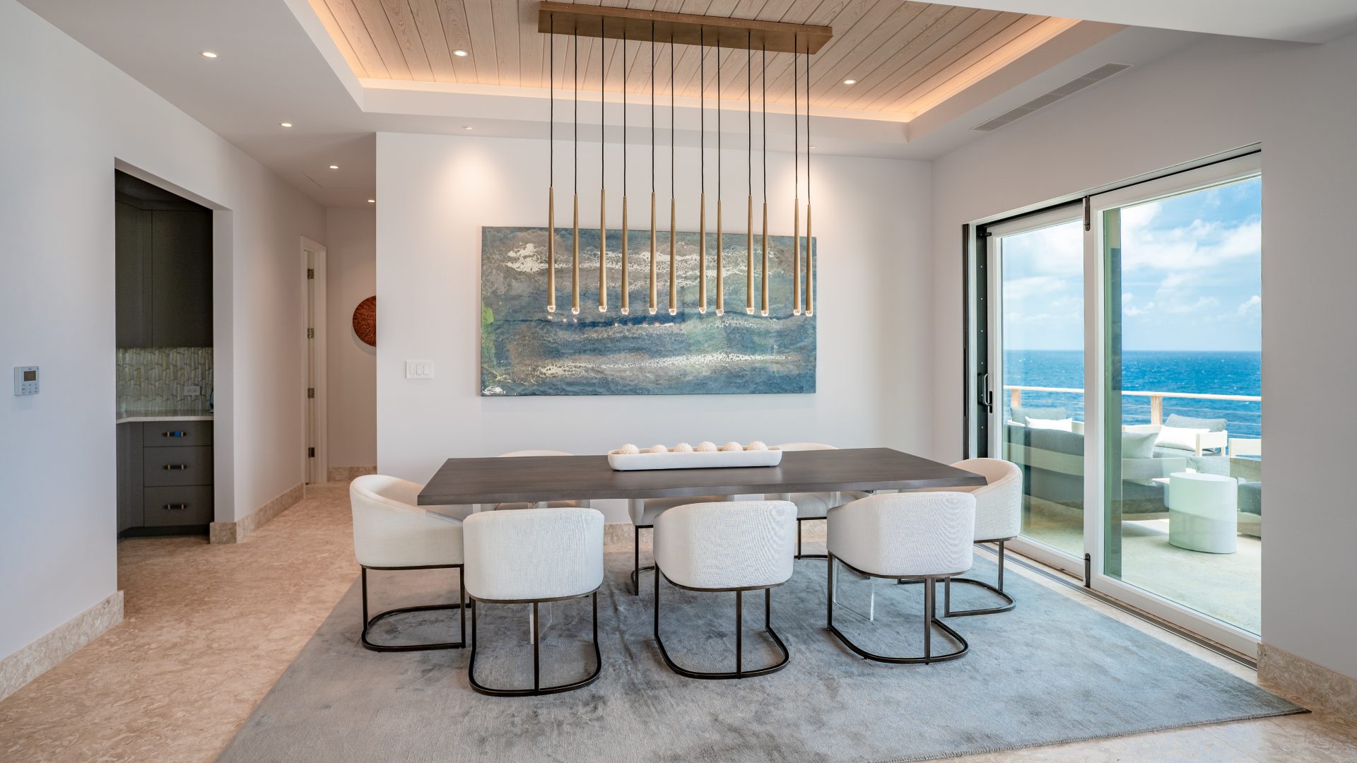 A modern dining room in the villa features a long table with eight white chairs arranged around it. Above the table hangs a linear chandelier with multiple pendant lights. A large, abstract painting decorates the wall, and sliding glass doors open to a view of the ocean where hummingbirds often visit.