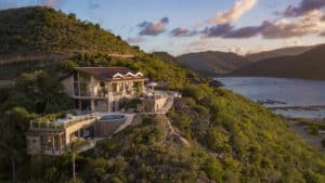 A luxurious cliffside villa with a pool, surrounded by lush greenery, overlooks a serene bay in the British Virgin Islands. The sky is painted with soft clouds during a warm sunset, and boats are visible in the water below. Rolling hills frame this picturesque scene.