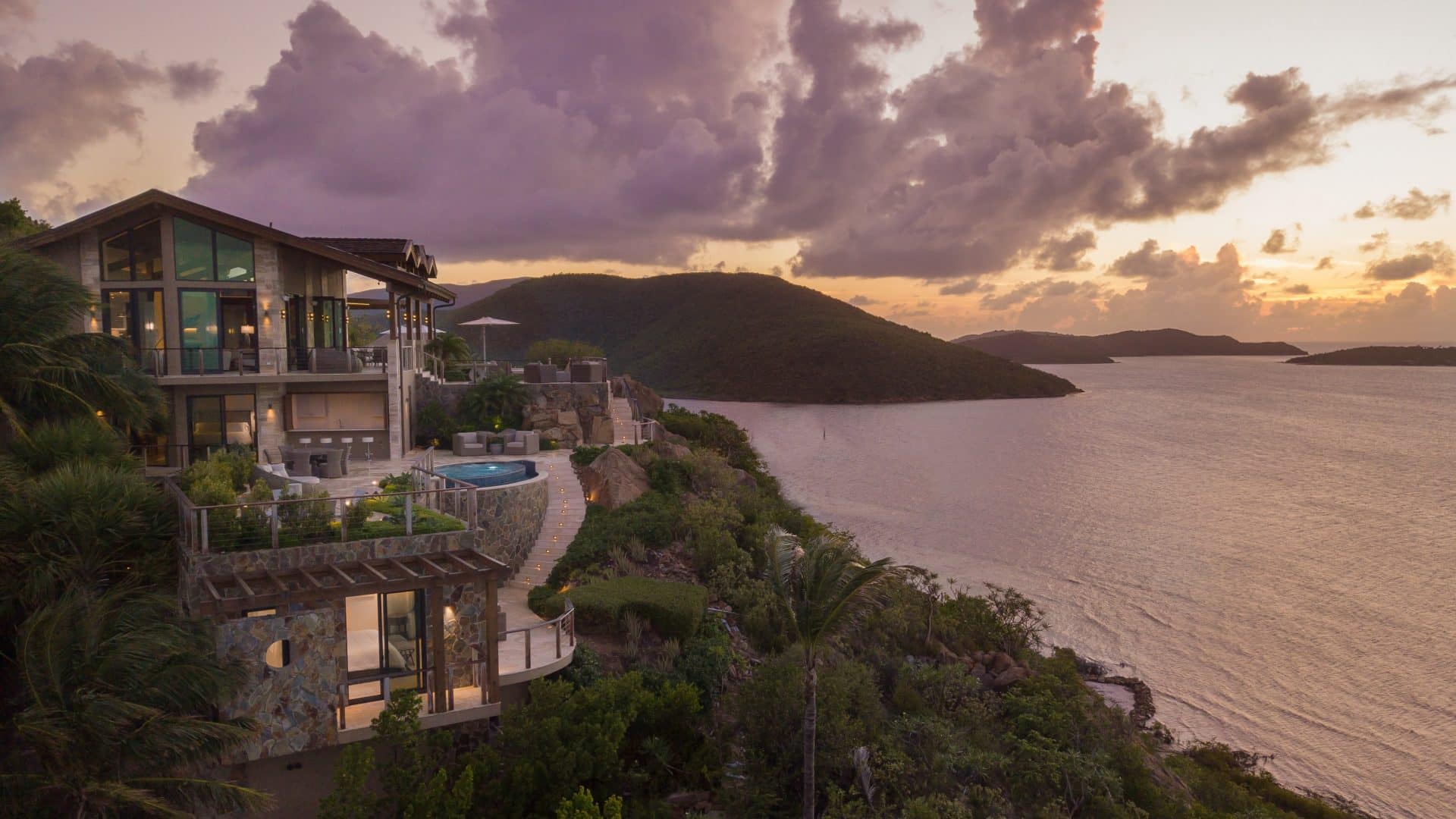 A luxurious multi-story house with large windows overlooks a serene body of water at sunset. The house features a pool and outdoor seating area on the cliffside, surrounded by lush greenery. The sky is filled with dramatic clouds and the scene is tranquil and picturesque—villas and homesites for sale in the BVI.