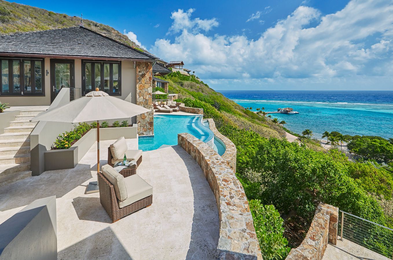 A luxurious outdoor patio at Oil Nut Bay features wicker furniture with cushions and large umbrellas beside an infinity pool overlooking a beautiful ocean view. Green hills and other villas are seen in the background under a partly cloudy sky, perfect for Discovery Tours enthusiasts.
