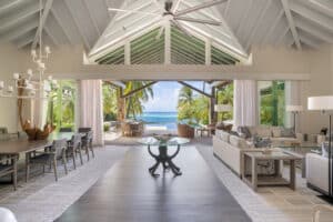 Spacious, modern open-concept living and dining area with high cathedral ceilings, large windows, and glass doors leading to a patio. The view extends to a tropical beach with palm trees and the ocean. At Nonamé Villa, a large round table sits in the center of the room.