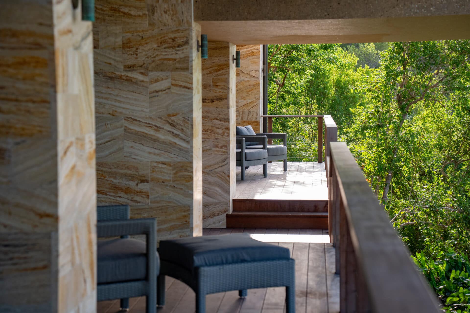 The sunlit balcony of Deep Bay Villa, featuring wicker furniture like a chair and ottoman, overlooks lush green trees. The stone-textured walls and wooden floor add to the serene ambiance of Villa DB1's natural setting.