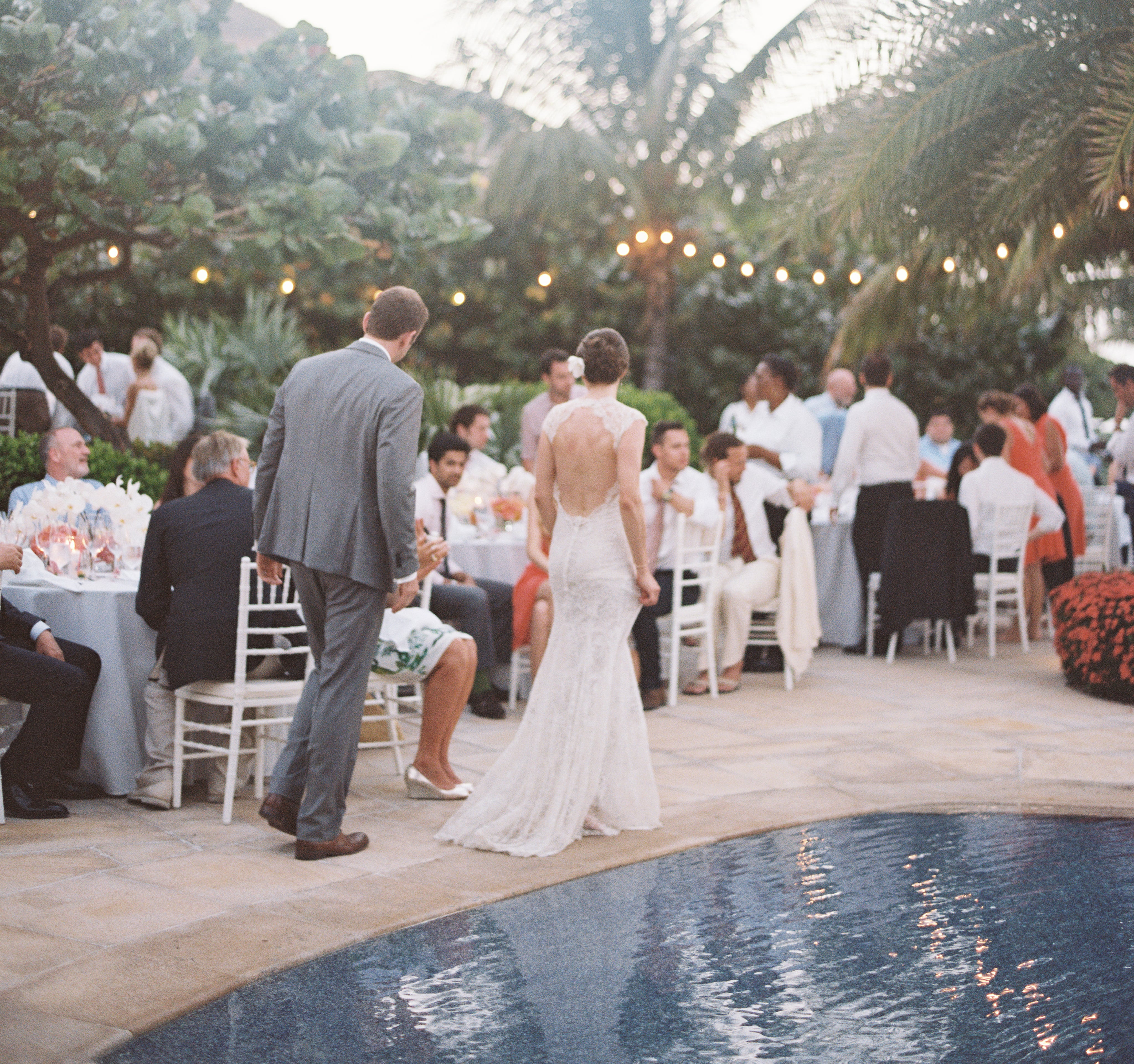 A bride in a white dress and a groom in a gray suit walk near a pool at an outdoor BVI Weddings & Events reception. Guests sit at round tables covered with white tablecloths, surrounded by greenery and trees adorned with string lights, creating a warm and festive ambiance.