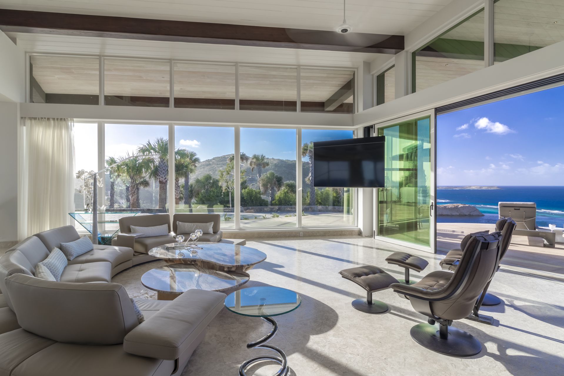 Discover a modern living room at Lion Heart Home in the British Virgin Islands, featuring large floor-to-ceiling windows offering ocean and palm tree views. Highlights include a curved sofa, round glass coffee table, armchairs, and wall-mounted TV. The outdoor patio with grill is visible through sliding glass doors. For sale now!