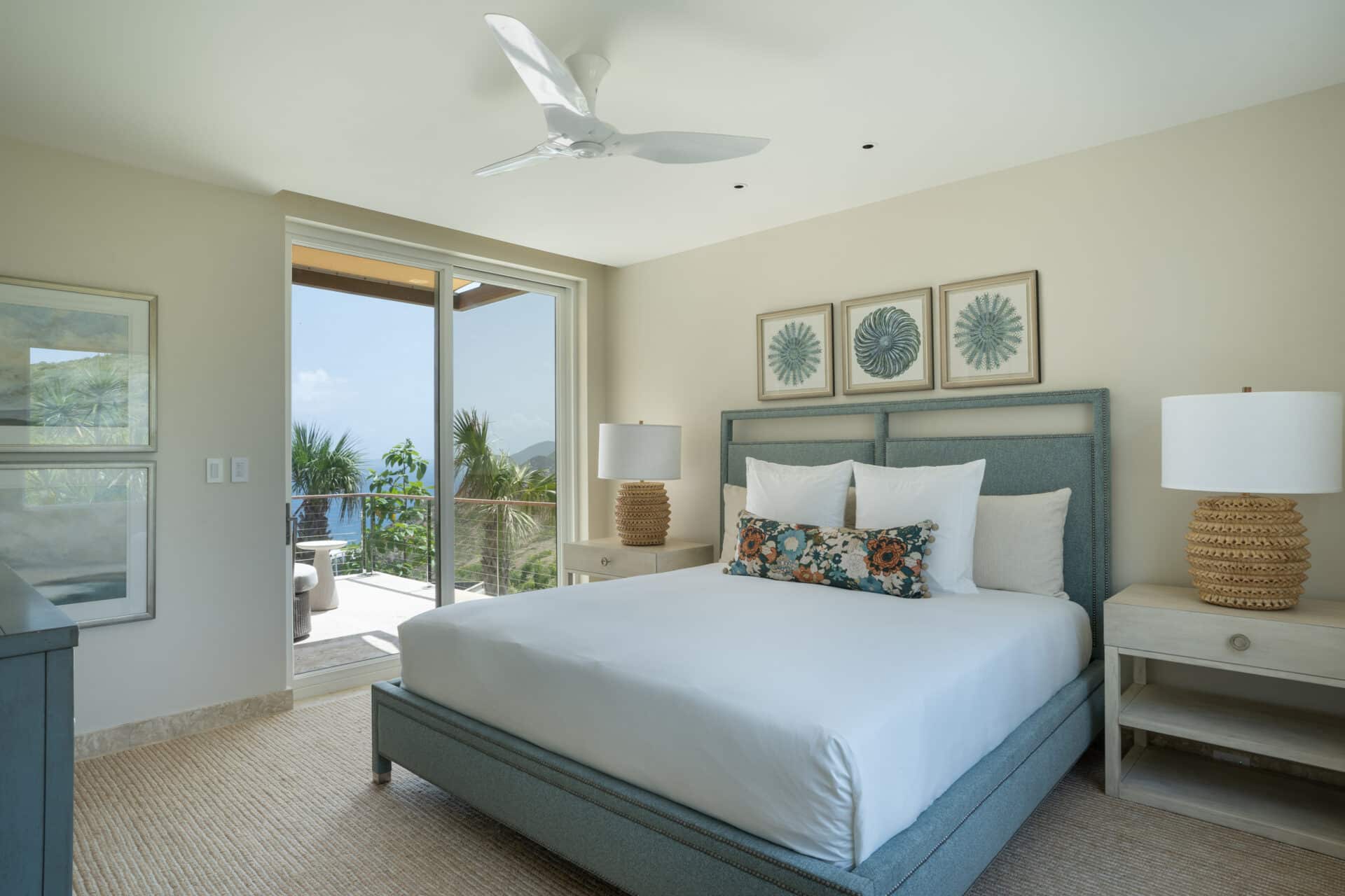 A serene Quintessa villa bedroom featuring a large bed with white linens and decorative pillows, flanked by two bedside tables with lamps. The room has light-colored walls, a ceiling fan, artwork above the bed, and a sliding glass door opening to a balcony with a scenic view.