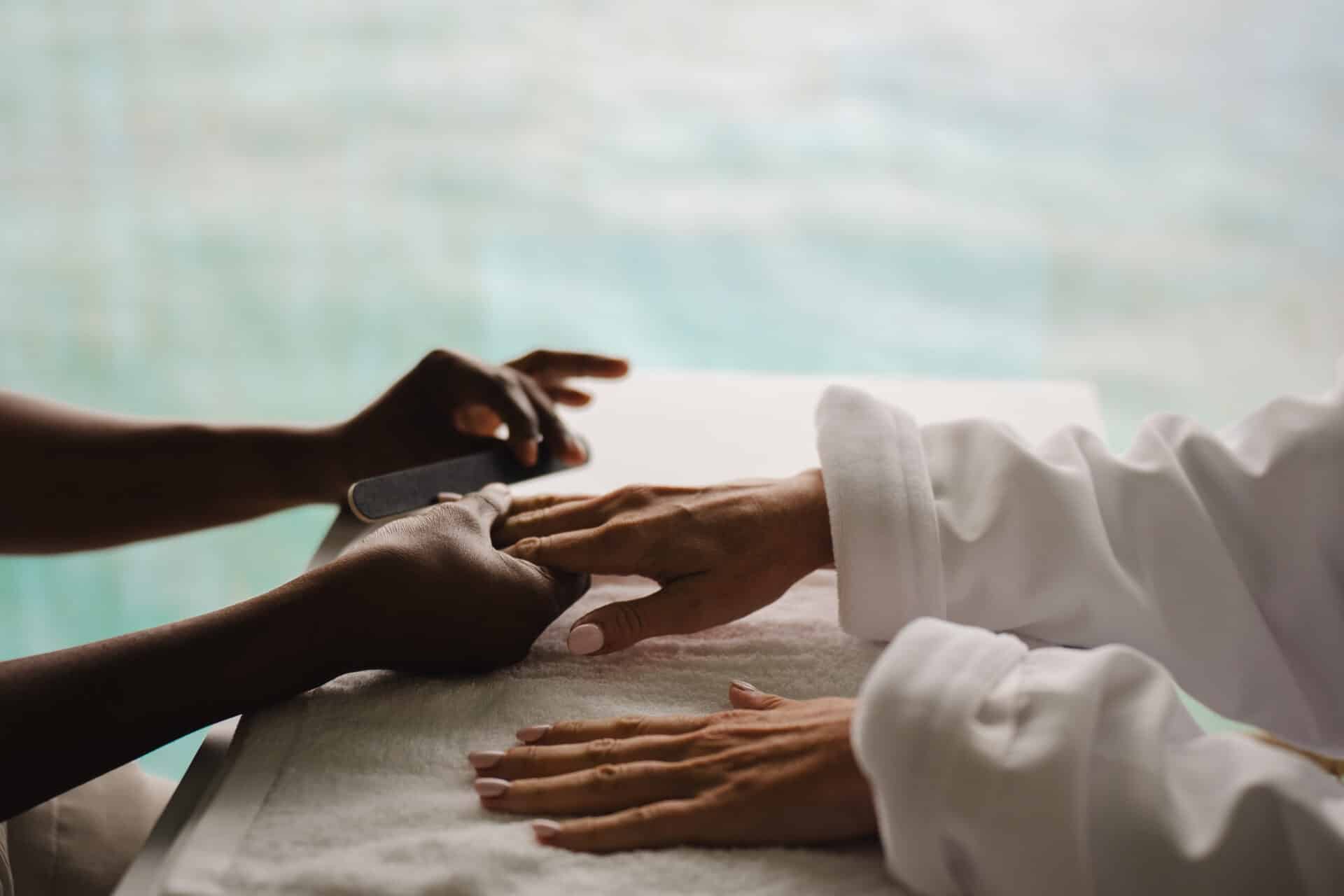 At Oil Nut Bay spa, a person in a white robe enjoys a manicure, their hands resting on a white towel as a nail technician expertly shapes their nails with a nail file.