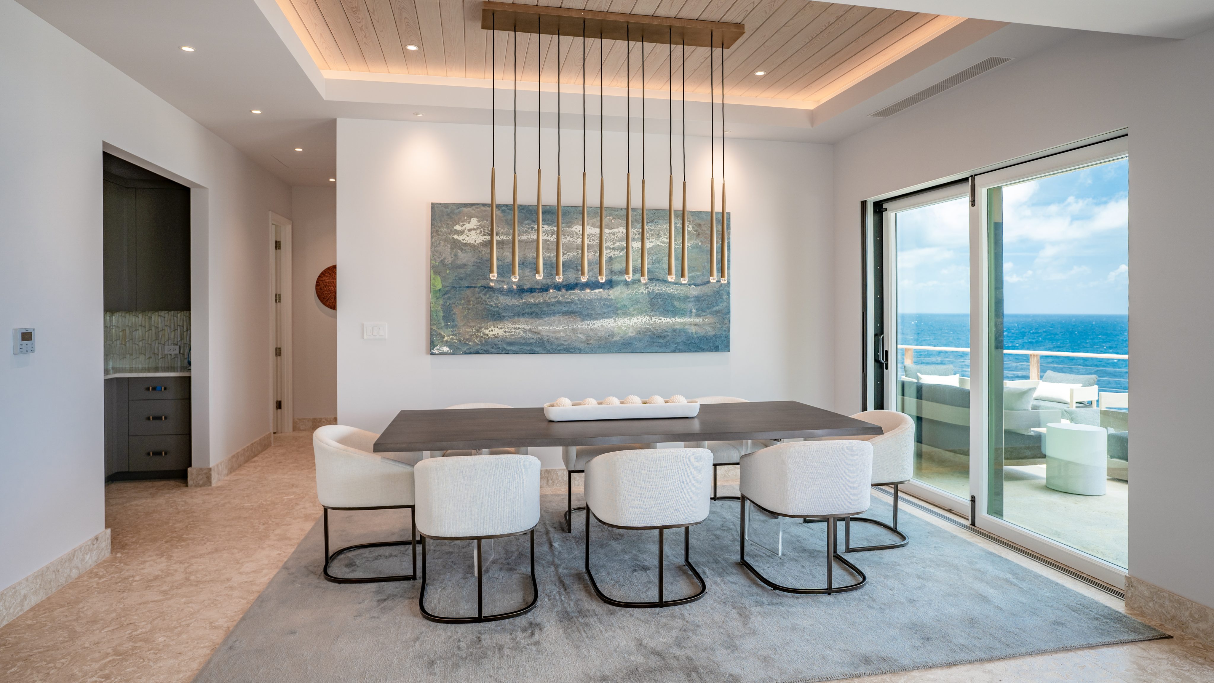 A modern dining room in the villa features a long table with eight white chairs arranged around it. Above the table hangs a linear chandelier with multiple pendant lights. A large, abstract painting decorates the wall, and sliding glass doors open to a view of the ocean where hummingbirds often visit.