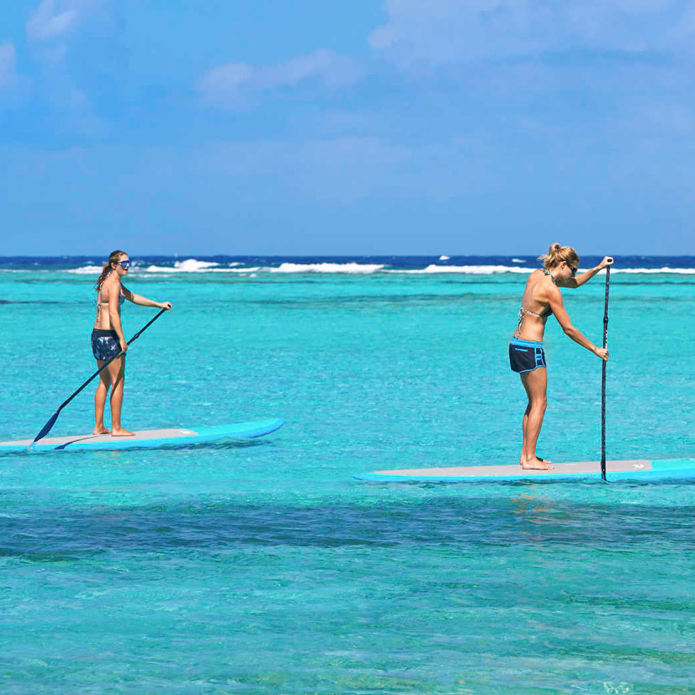 Two people paddleboarding on clear turquoise water at Oil Nut Bay. Both are standing on separate boards in swimwear, using long paddles to navigate. The horizon in the background shows a deep blue ocean and some light waves, with a bright blue sky above.