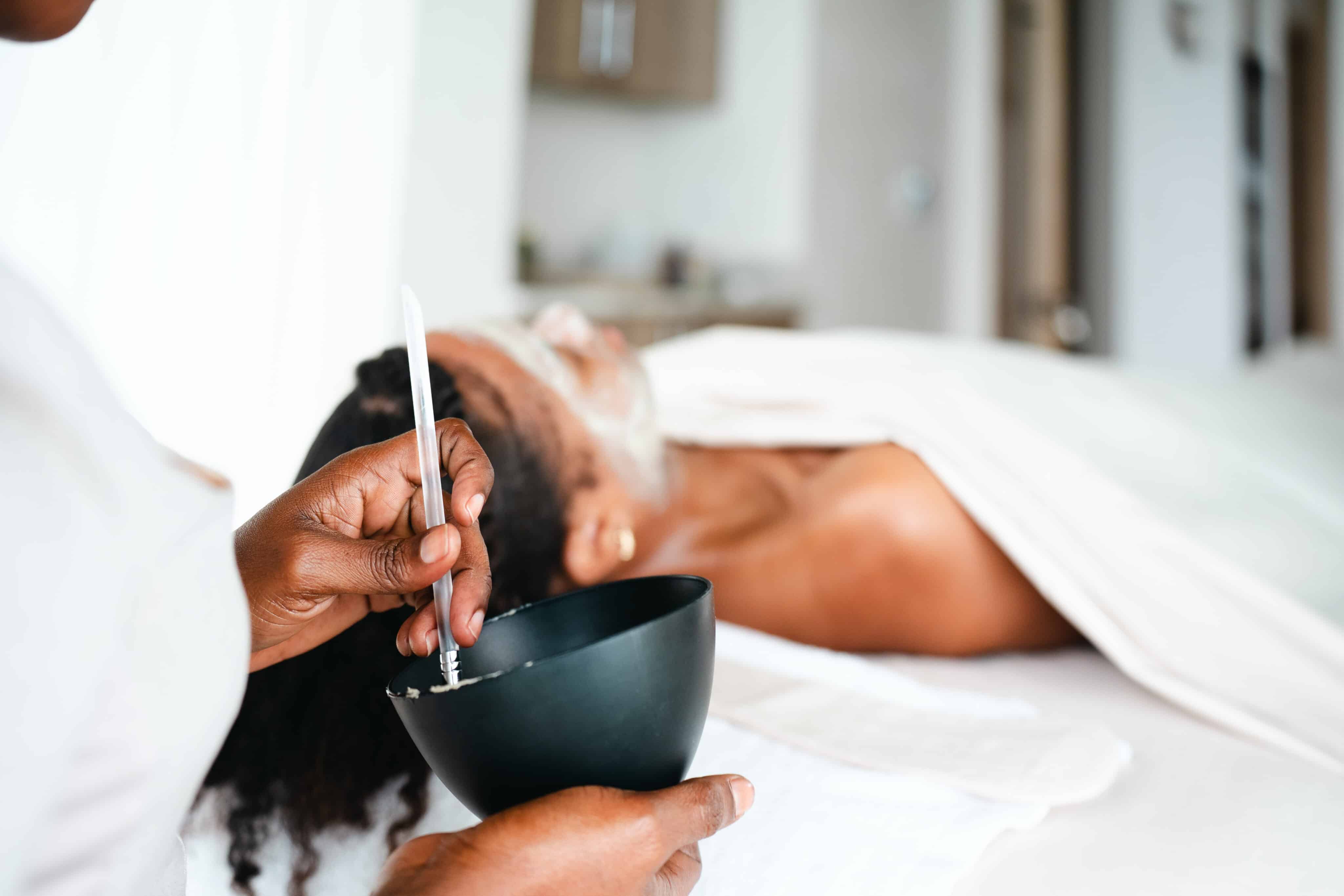 A person lies on a spa bed with a white towel, receiving a soothing facial at Oil Nut Bay spa. Another attendant mixes a facial mask in a black bowl, applying it gently in the calm and relaxing setting.
