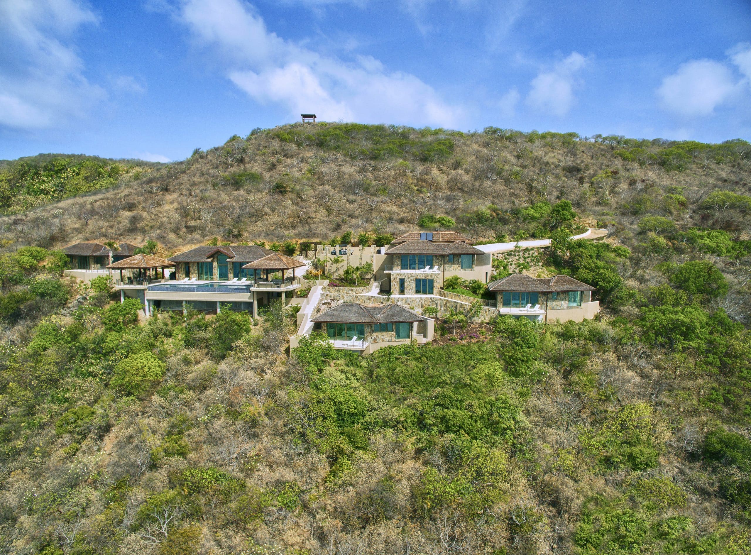 A luxury villa complex is nestled on a lush, mountainous landscape under a clear blue sky. The interconnected buildings, resembling an elegant reef of houses with green roofs and large windows, are surrounded by greenery and connected by a winding pathway.