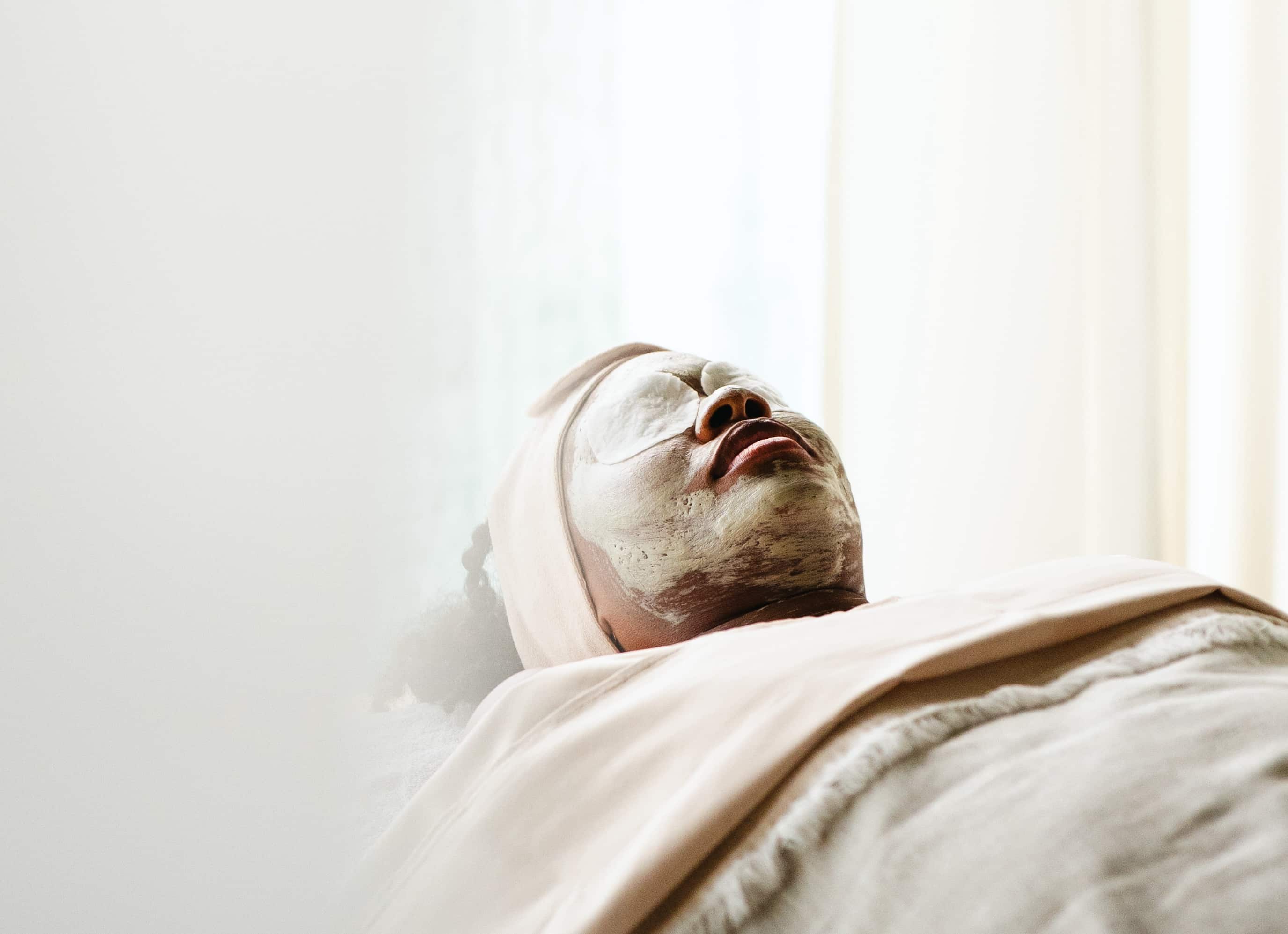 A person is lying down with eyes closed, wearing a headband and facial mask at Oil Nut Bay spa. They are covered with a beige blanket, relaxing in a softly lit, serene spa environment.