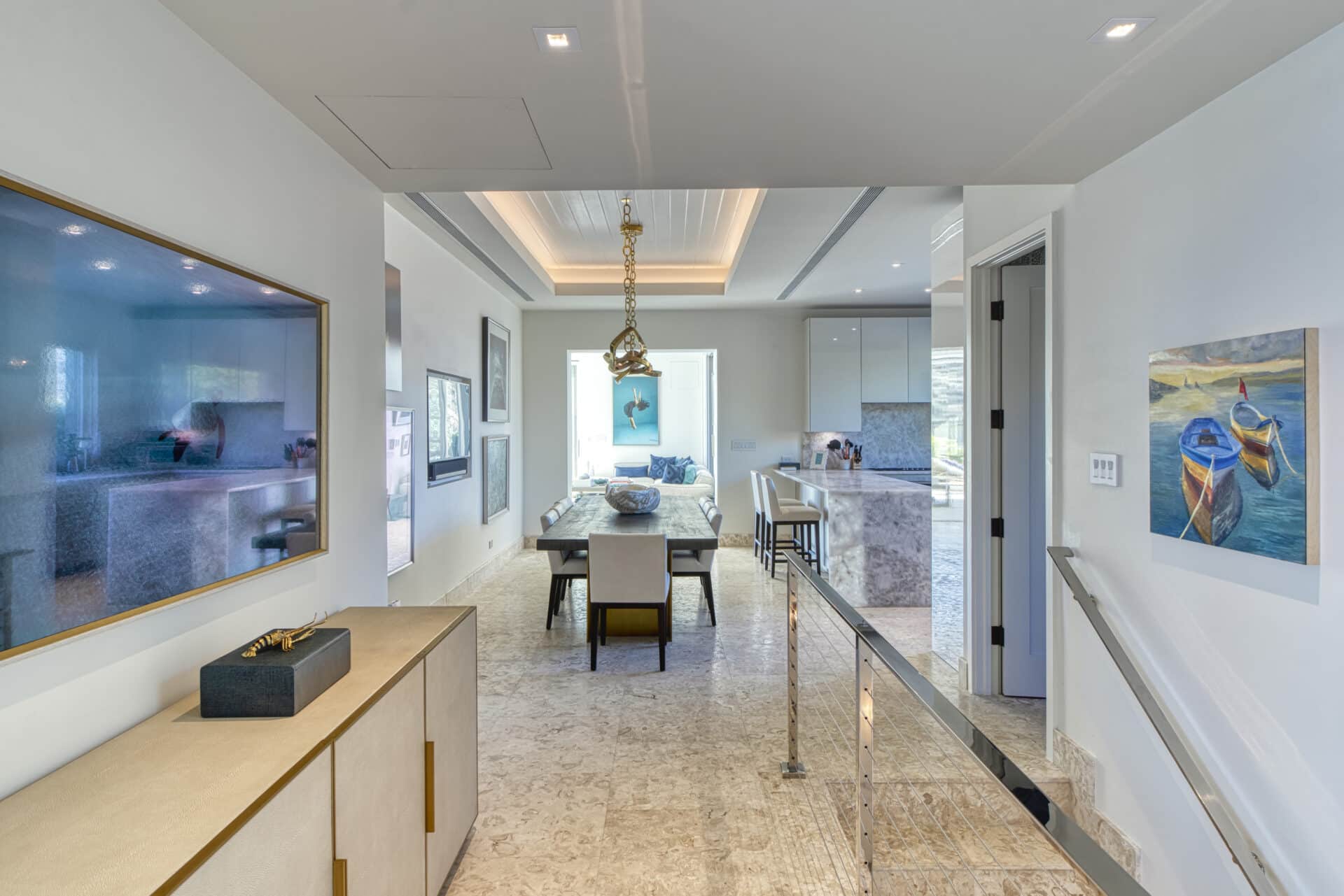 The modern dining room at Wild Side Villa boasts a rectangular dining table with eight chairs. It features a large photo on the left wall and a framed painting of two boats on the right. The space is completed with a light-colored marble floor and recessed lighting in the ceiling.